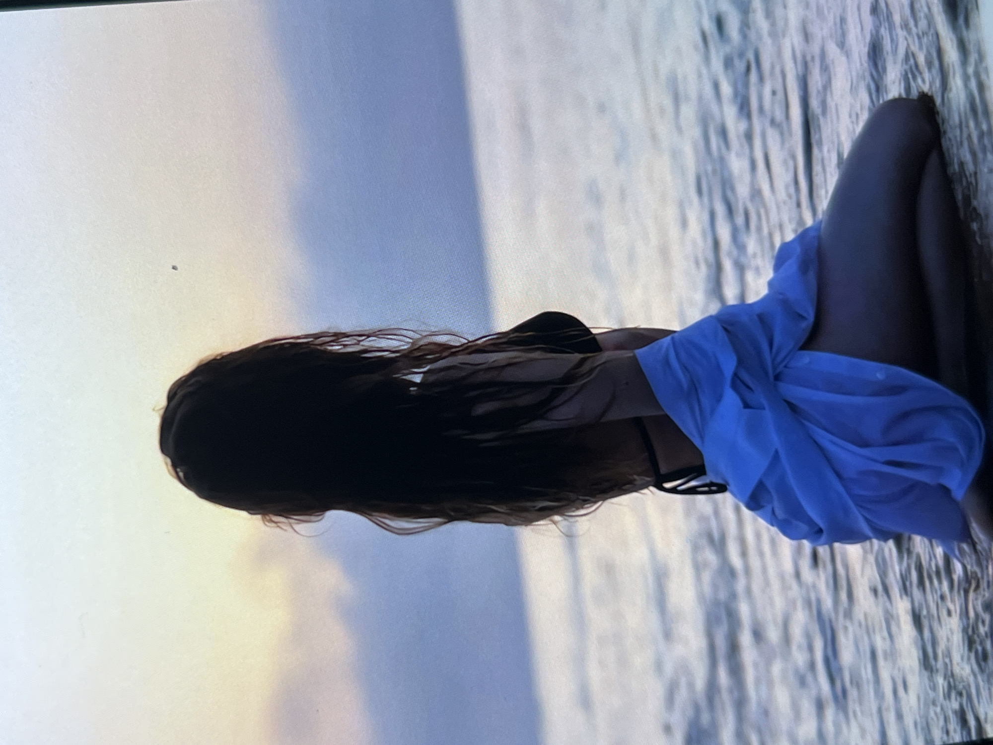 A person with long, dark, wet hair is kneeling in shallow water at the edge of a body of water, possibly the sea or a large lake. They are facing away from the camera, looking towards the horizon where the sky transitions from a pale yellow near the horizon to a muted blue above. The person is wearing a black bikini top and a light blue garment wrapped around their lower body. The water is calm with gentle ripples. The overall scene suggests a serene, possibly sunset or sunrise atmosphere. The location is not specified in the image and cannot be determined from visual cues.