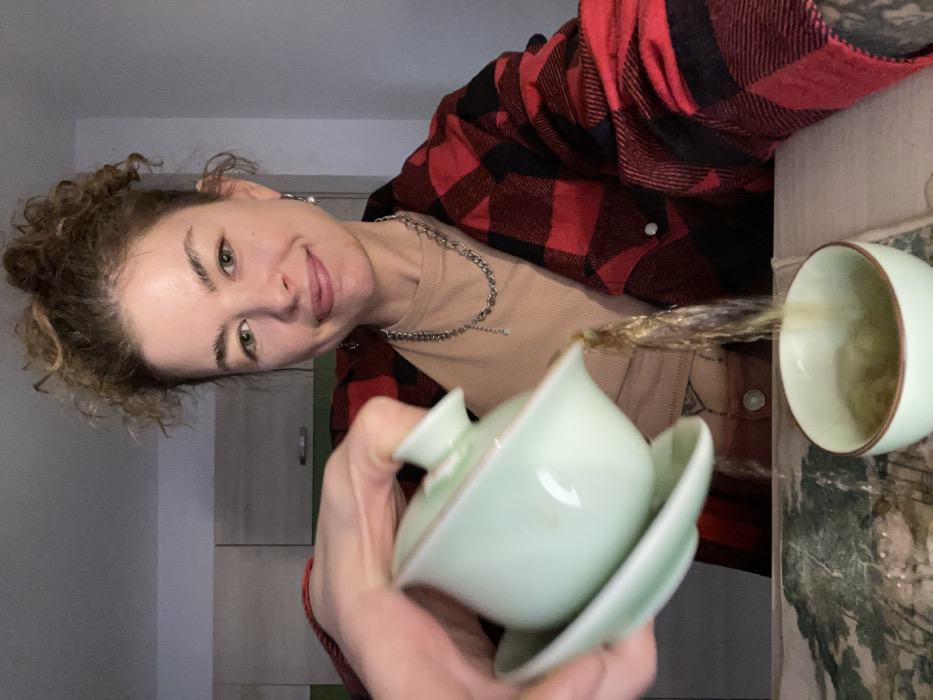 A woman in a red and black flannel smiles sideways at the camera while pouring tea from a pale green lidded cup into a matching teacup. The tea forms a golden stream mid-pour. The woman wears a silver chain necklace and a neutral-colored shirt beneath the flannel. The background shows a hint of a cabinet and a surface with a decorated mat beneath the teacup.
