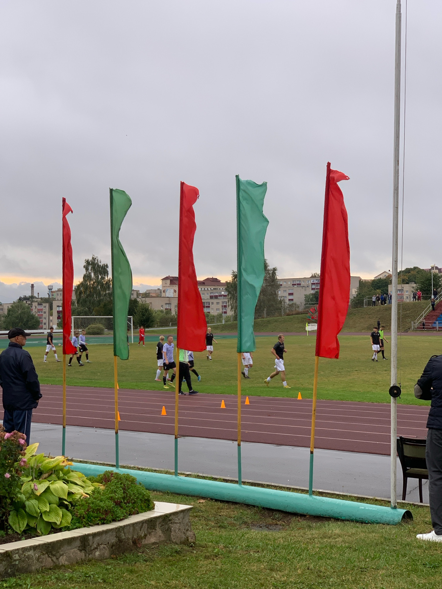This image depicts an outdoor football (soccer) match on a cloudy day in Kapyĺ, Belarus. The scene is set on a well-maintained green football pitch with a running track surrounding it. In the foreground, there are several tall, brightly colored flags – two red and one green – mounted on poles, creating a visual barrier. To the left, a person in a dark jacket and cap stands observing the game.
On the football pitch, two teams of young male players, distinguishable by their uniforms (primarily white and black with some accents), are actively engaged in the game. Several players are in motion, suggesting the match is in progress. One player in black and white is running with the ball, while others are positioned on the field, prepared to receive passes or defend. A goalpost is visible in the background, with a player in a red uniform near it.
Behind the pitch, residential buildings and some trees are visible, indicating an urban or semi-urban setting. A stadium stand with seating is also present in the far right of the image. The sky is overcast, suggesting a potentially cool or damp day, and the lighting indicates it is likely late afternoon. The overall mood appears to be one of focused sporting activity, with the flags adding a festive or ceremonial touch to the event. No visible text is discernible in the image.