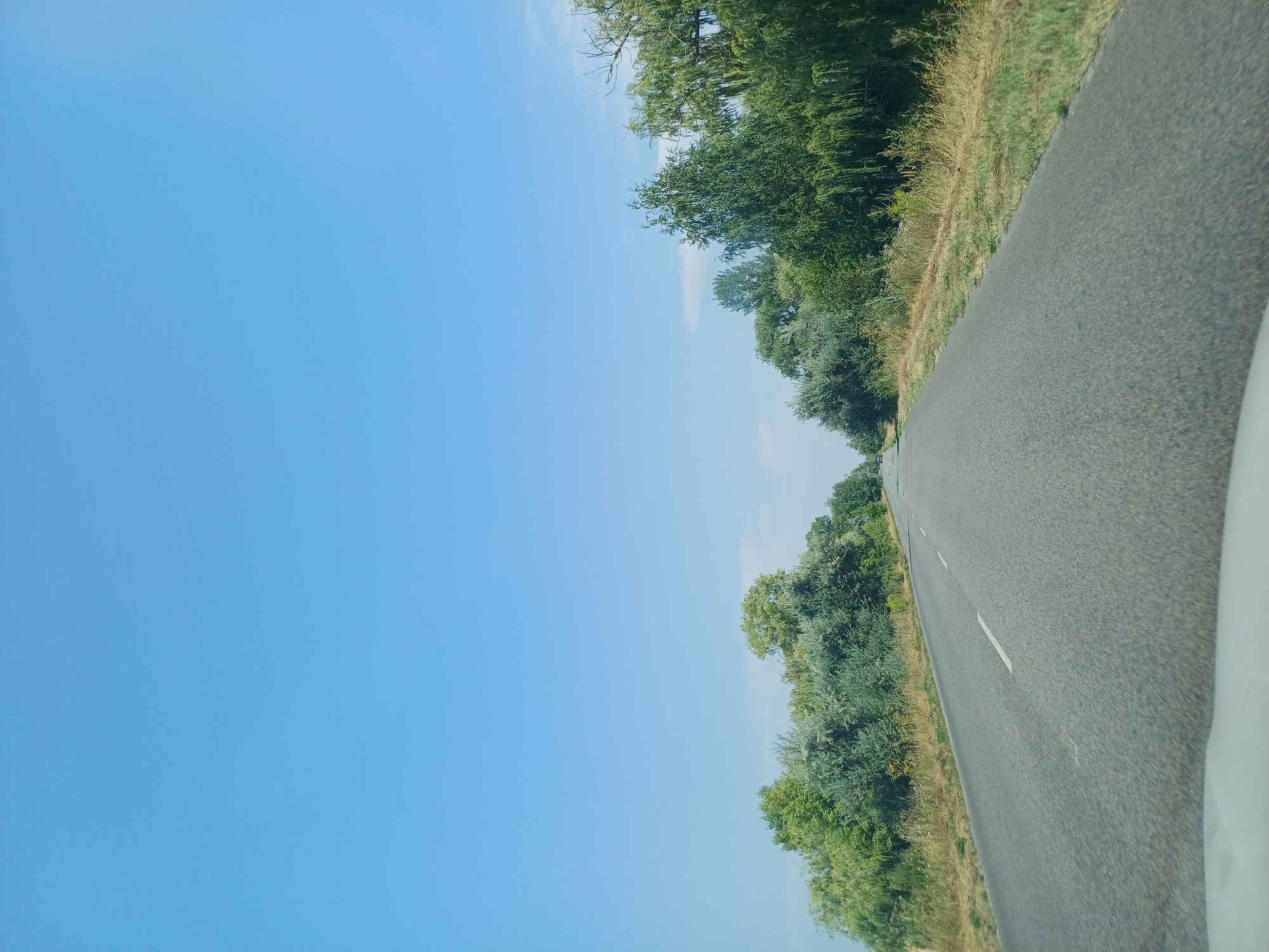 A clear, bright blue sky dominates the upper portion of the image. Below the sky, a paved road stretches into the distance, flanked by lush green trees and vegetation on both sides. The road has a white dashed line marking the center. The scene appears to be a rural or semi-rural area, possibly on the outskirts of Boryspil', Ukraine, given the natural landscape. There are no people or vehicles visible in the immediate foreground, but a distant car can be seen on the road in the far left. The overall impression is one of a peaceful, sunny day on a country road.