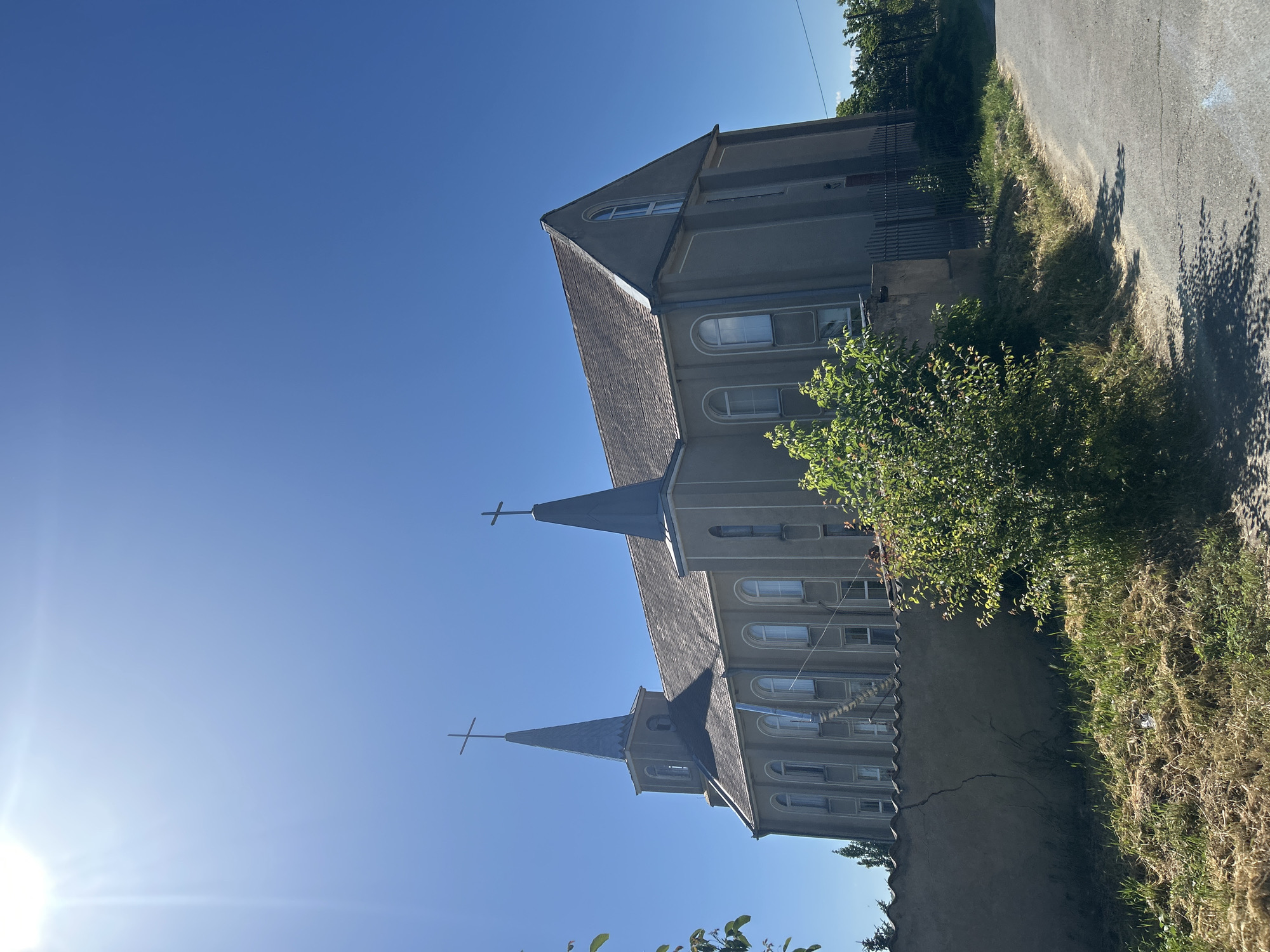 This is a vertical, wide-angle photograph taken on a sunny day in Ivanivka, Ukraine. The main subject is a church with two spires, each topped with a cross. The church is made of gray material and has a gray, sloped roof. Many arched windows are visible on the side of the building. A bright sun is visible in the lower left corner of the image, casting a lens flare. To the right of the church is a grassy area with some dry grass and a small tree. A paved road is visible in the upper right portion of the image, with shadows of trees cast across it. The sky is a clear, bright blue. There is no visible text in the image.