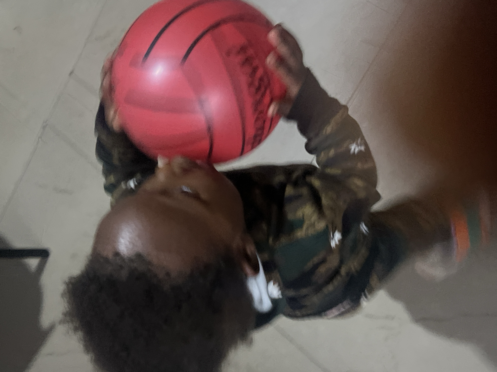 A child is holding a red basketball. The environment appears to be indoors with a light-colored tile floor. The child has dark curly hair and is wearing a camouflage patterned shirt. The location is assumed to be Gura Top, Nigeria.