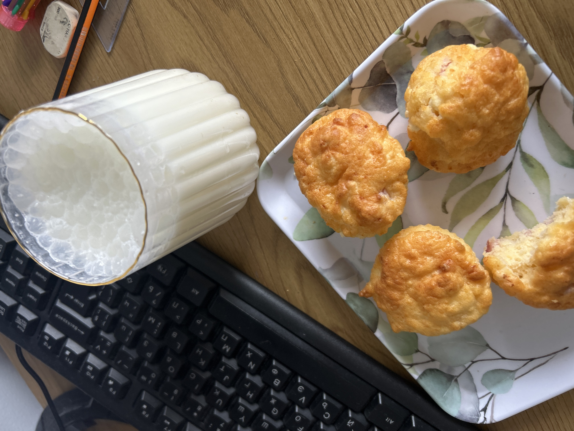 A top-down shot shows a desktop scene. On the left, a black computer keyboard is positioned next to a white candle in a ribbed glass holder with a gold trim. To the right of the candle, a square plate with a floral leaf pattern holds four muffins. One of the muffins has a bite taken out of it. In the upper left corner, a collection of office supplies including colored pencils, an eraser, and a ruler are visible. The surface is a light brown, possibly wood. The location is not discernible without further information.