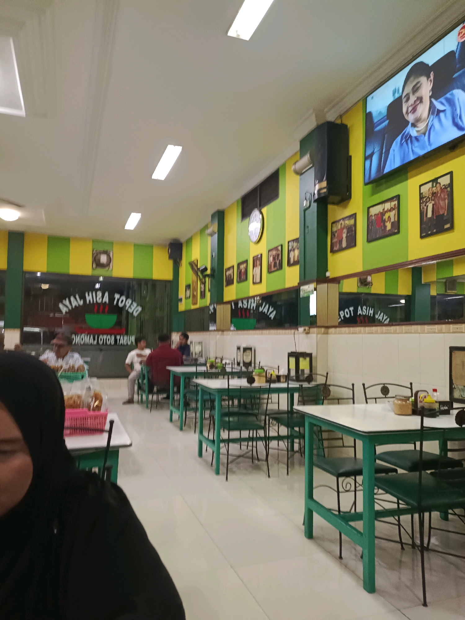 The image depicts the interior of a restaurant in Dapur, Indonesia. The restaurant has a vibrant aesthetic with yellow and green striped walls. A large television screen displays a smiling woman in a blue shirt. Framed photographs are mounted on the walls, and a clock is visible. Tables and chairs, predominantly green, are arranged throughout the dining area. Some patrons are seated at the tables, engaged in dining. The flooring appears to be tiled. A large mirror reflects some of the interior. The overall atmosphere suggests a busy eatery.