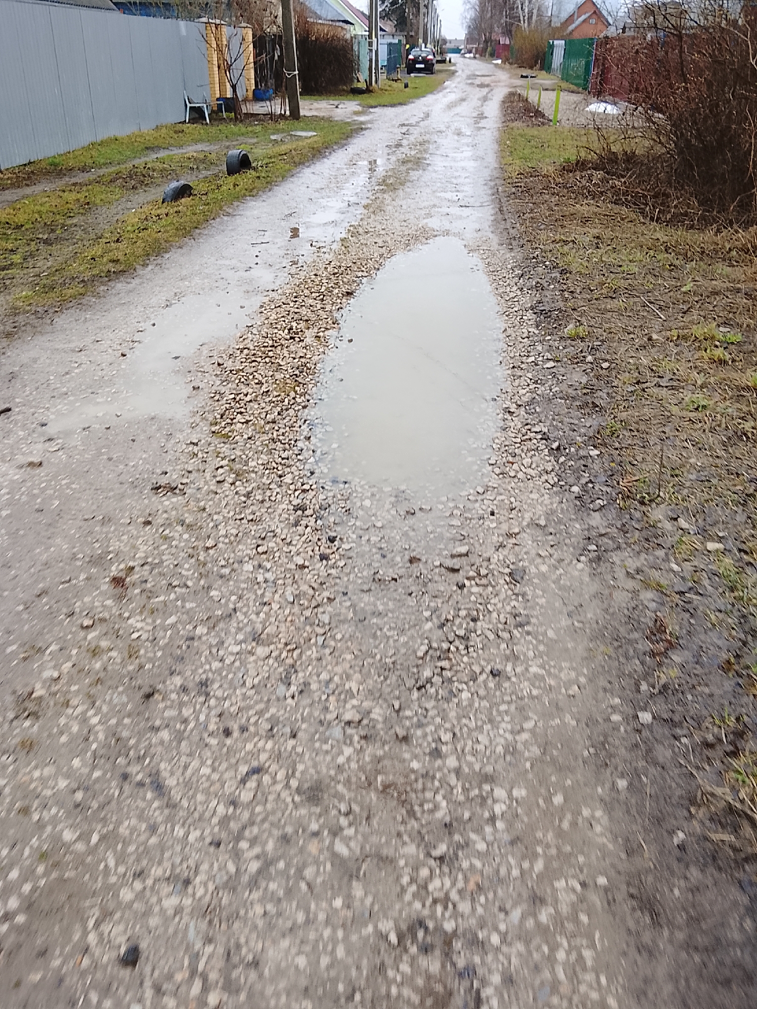 The image depicts a waterlogged dirt road in what appears to be a residential area in Высокиничи, Russia. The road surface is primarily gravel and dirt, with several puddles of varying sizes. Two black tires are visible on the grassy verge to the left of the road. In the background, houses and fences are visible, along with utility poles. The scene suggests a rural or semi-rural environment, possibly after recent rainfall.