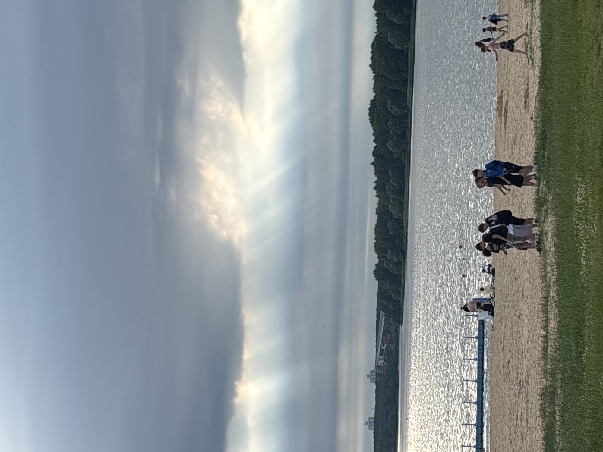 The image depicts a daytime scene at the Minsk Sea, a reservoir located on the Svislach River in the western part of Minsk, Belarus. The sky is overcast with dramatic sunbeams breaking through the clouds, creating a dappled effect over the water. In the foreground, a sandy beach is visible with several groups of people.
On the beach, there are at least ten individuals, appearing to be mostly young males, some walking, others standing in small groups. One group of four boys is walking towards the right of the frame, dressed in casual clothing, including shorts and t-shirts. Another group of three boys is walking from left to right in the center of the image, one wearing a blue jacket. Further to the left, near the water's edge, a small group of people is gathered around some bags, appearing to be interacting. A blue floating platform is positioned in the water near the beach.
The water is choppy and reflects the light from the sky. Beyond the water, a dense line of green trees forms the background, with some distant buildings and a bridge visible on the horizon, indicating an urban or suburban setting. The overall atmosphere is that of a recreational area on a cloudy but bright day.