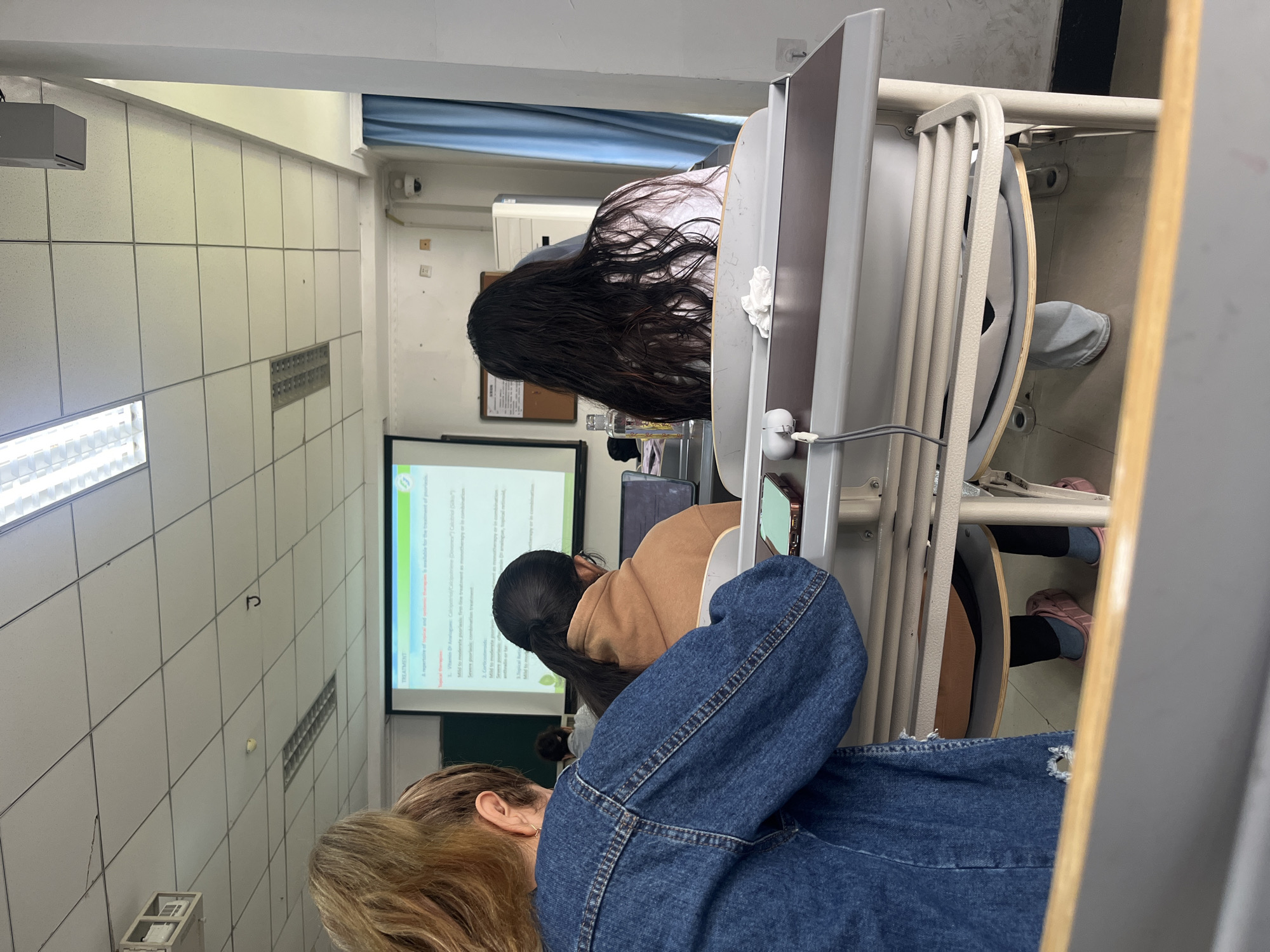 This rotated image captures an indoor classroom or lecture hall from the perspective of someone seated, looking towards the front. Several individuals are visible, seated at desks and chairs, all facing forward towards a projected presentation.
In the immediate foreground, the back of a person with blonde hair, wearing a blue denim jacket with distressed elements, occupies the lower left corner. Behind them, another person with dark hair tied in a ponytail, dressed in a brown jacket, is seated at a desk. Further back, on the right side of the frame, is a person with long, dark, wavy hair, wearing a light-colored top, also facing the front. Personal items like a water bottle, a mobile phone, and a crumpled tissue are visible on their desk.
The classroom features white tiled walls with visible grout lines and a rectangular vent. A fluorescent light fixture runs along the upper left ceiling, and a projector is mounted above, casting an image onto a large white screen at the front of the room. A surveillance camera is mounted high on the right wall. The feet of some attendees are visible beneath the desks, with some wearing pink footwear.
The projection screen displays a slide with "TREATMENT" as the main heading. Visible text snippets include "Mild to moderate psoriasis: first-line treatment as monotherapy or in combination," "Severe psoriasis: combination treatment," and "2. Corticosteroids:". There are also mentions of "Topical Therapies:" and "1. Vitamin D3 Analogue: Calcipotriene/Calcipotriol (Dovonex)/Calcitriol (Silkis)". The content suggests a medical or scientific lecture. The scene depicts an educational session with artificial lighting.