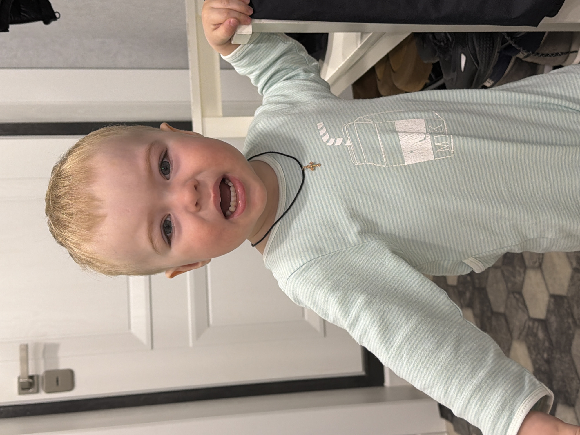 A young child, with fair skin and blonde hair, is smiling with an open mouth. The child is wearing a light blue and white striped long-sleeved shirt with a graphic of a glass container on the front and a necklace with a cross pendant. The child is holding onto the edge of a white shelf. The shelf contains shoes. A white door with a black frame and a silver door handle is visible. Part of the floor has a hexagonal pattern. It cannot be determined from the image if the photo was taken in Votkinsk, Russia.
