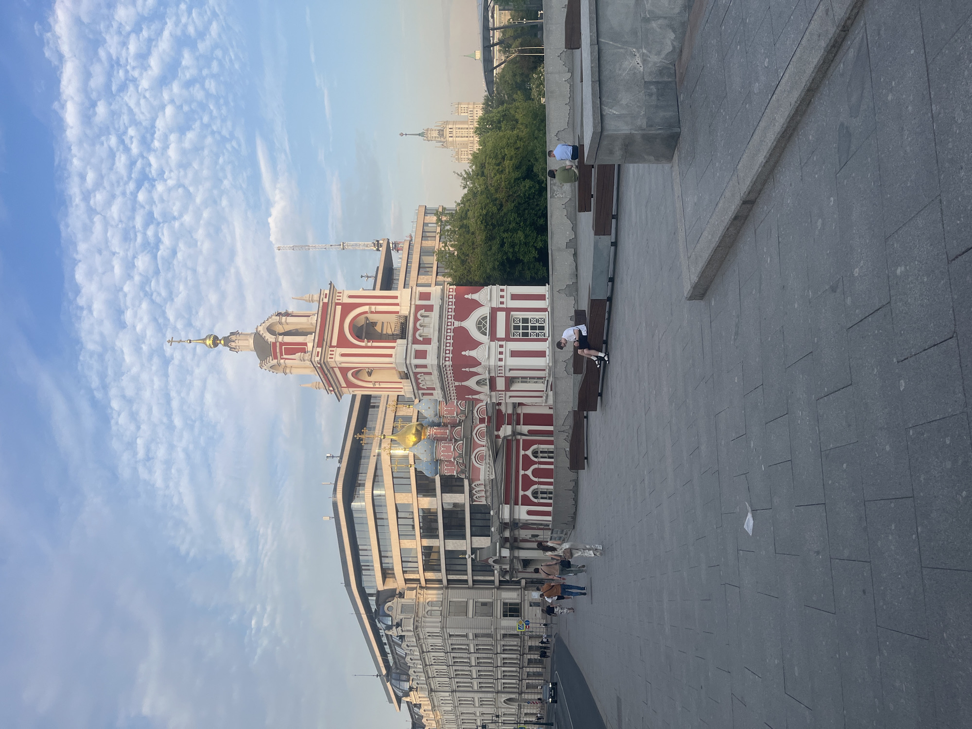 The image depicts a street scene in Moscow, Russia, featuring a prominent red and white church with a golden dome, juxtaposed against modern architecture. Several people are visible walking and sitting on benches along a paved walkway. In the background, a tall, Stalinist-era skyscraper stands out against the sky, surrounded by trees. The sky is partly cloudy, with a mix of blue sky and white clouds. The overall impression is a blend of historical and contemporary urban elements.