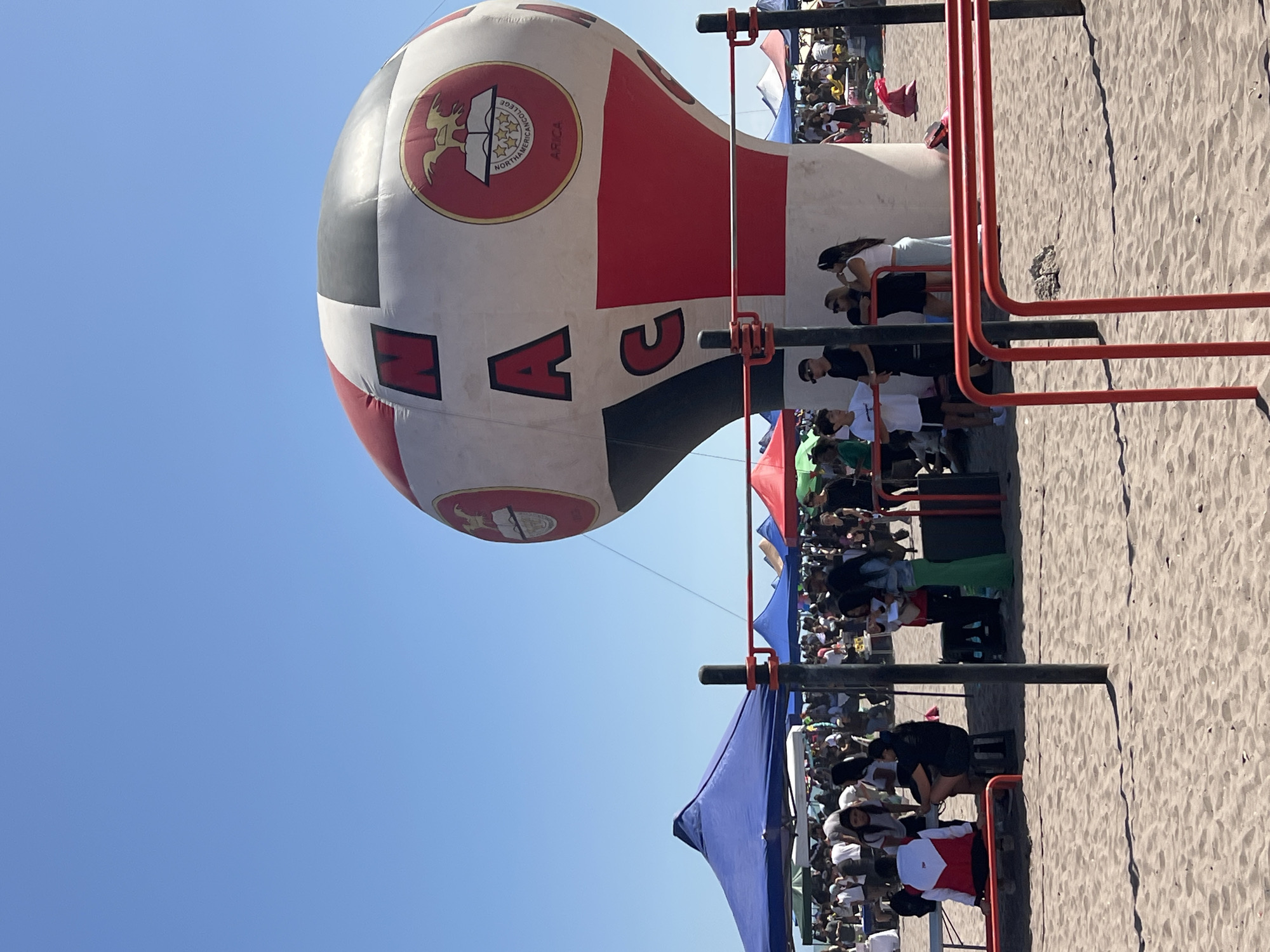 The image depicts a crowded beach scene, likely in Arica, Chile, as indicated by the "North American College Arica" logo on a large, egg-shaped balloon or inflatable structure. The balloon has a white background with red and black accents, prominently displaying the letters "N A C". Below the balloon and to the right, a dense crowd of people is visible, standing on the sandy beach. Red and blue tent canopies partially cover the crowd. In the foreground, red metal poles or structures are visible, possibly part of a barrier or railing. The clear, blue sky occupies the upper left portion of the image. The overall scene suggests a public event or gathering near the beach in Arica.