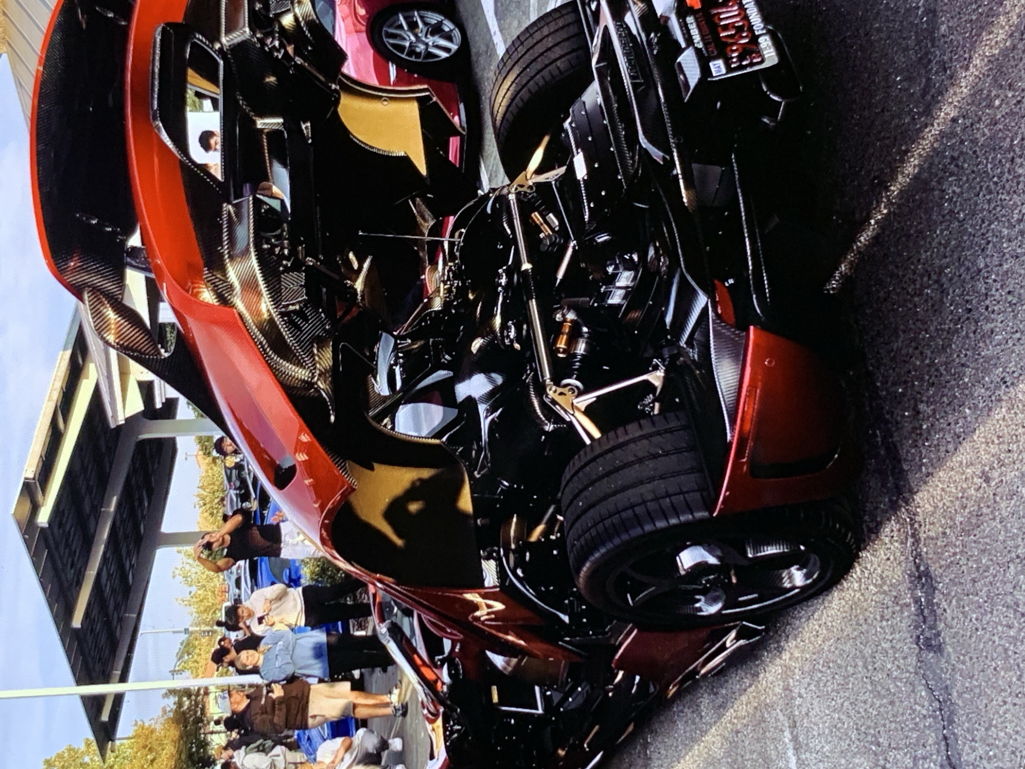 The image depicts an outdoor gathering, possibly a car show or meet-up, in an urban environment. In the foreground, a red sports car with its hood and engine bay open is prominently featured. The car's intricate mechanical components and carbon fiber accents are visible. Several people are gathered around, some appearing to be spectators, while others might be owners or enthusiasts. One person is seen taking a photo with their phone. In the background, other vehicles, including a red sports car, are parked, and a structure with a roof, possibly a parking garage or outdoor shelter, is visible. The ground appears to be asphalt. The overall scene suggests a casual event with a focus on automotive appreciation.