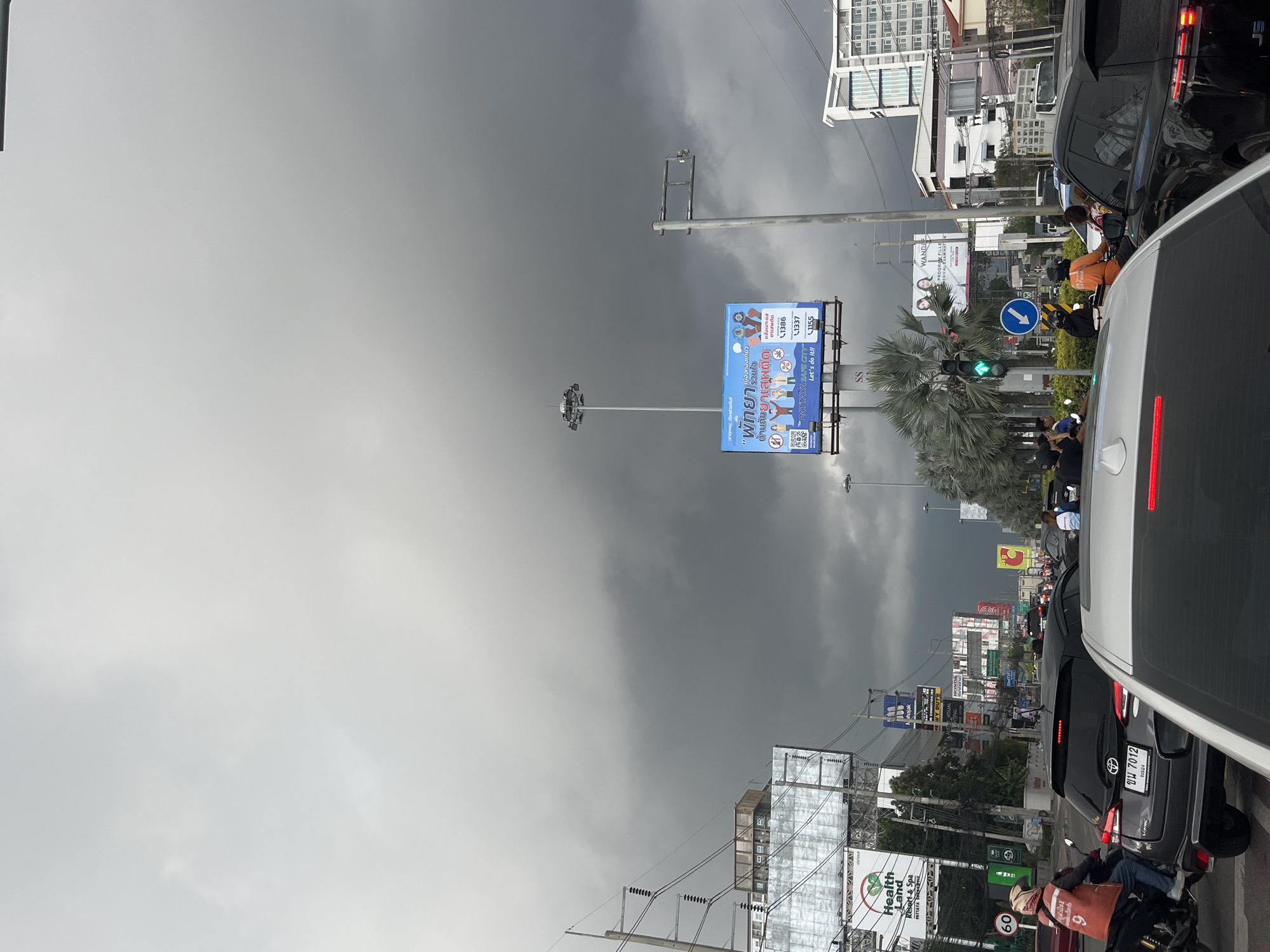 The image depicts a street scene in Pattaya City, Thailand under a cloudy sky. Traffic is present, with cars visible including a silver car and a dark grey Toyota Vios with the license plate ขน 7012. A motorcycle rider wearing an orange uniform and a mask is also visible near the Health Land Resort & Spa sign. The road is lined with various billboards and signs, including an anti-drug campaign banner and advertisements for Health Land Resort & Spa. Traffic lights are seen with a green light. Palm trees and electrical wires are present. Buildings and other structures line the street, and a tall lamppost stands along the street.