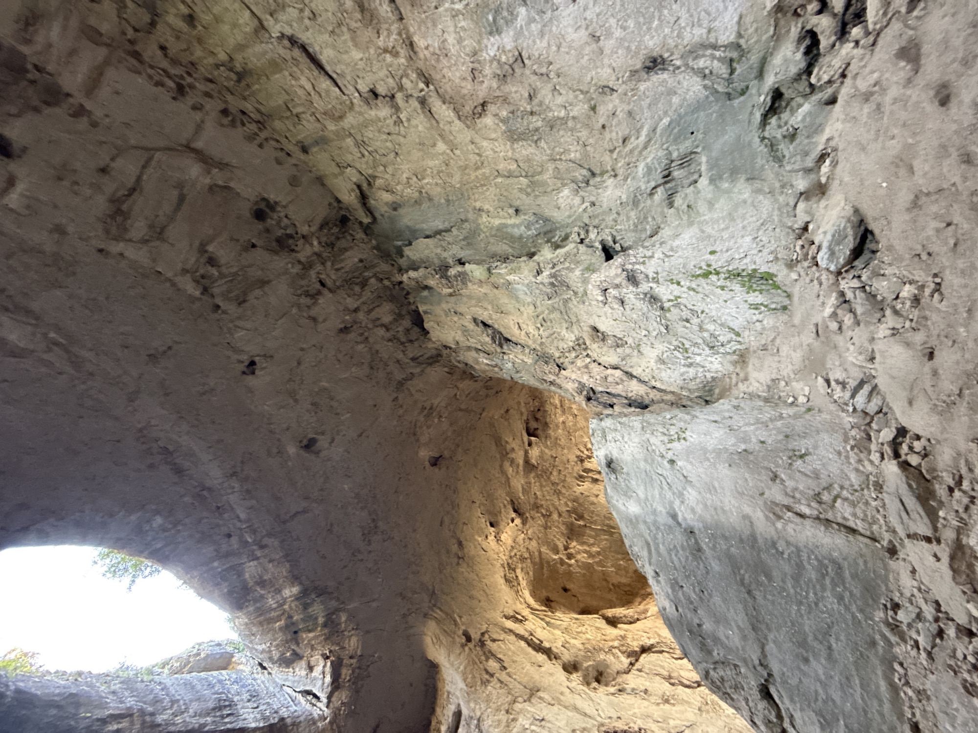The image depicts the interior of a large cave, likely a natural rock formation. The cave ceiling is a prominent feature, appearing smooth and arched, with light filtering in from an opening at the top. The walls are rough and textured, displaying layers of rock and numerous small holes or indentations. Large boulders and rock debris are scattered across the cave floor, suggesting a natural and undeveloped environment. The lighting suggests it is daytime, with sunlight illuminating parts of the cave's interior, creating areas of shadow and highlighting the rock formations. The scene is devoid of any human presence or activity. This imagery is consistent with the geological features found in the karst region of Karlukovo, Bulgaria, known for its extensive cave systems and natural arches, such as the "Eyes of God" rock formation.