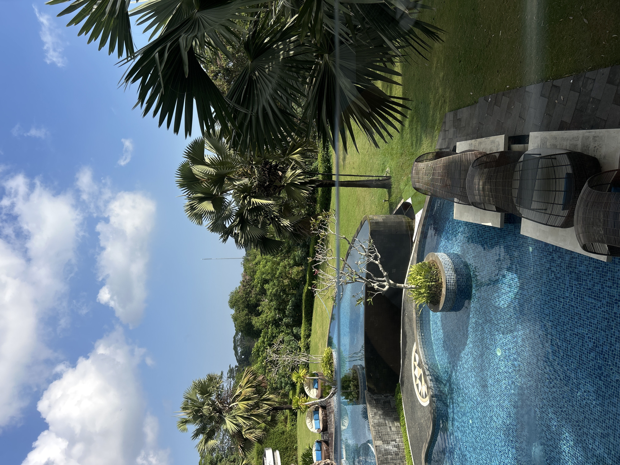 The image shows an aerial view of a swimming pool area with lush green landscaping and palm trees. The pool is blue and tiled, with several circular islands featuring small trees and plants. There are several woven lounge chairs with blue cushions placed on a paved area beside the pool. In the background, there is a dense green forest and a clear blue sky with white clouds. The scene appears to be a resort or a well-maintained private property in Kuta Selatan, Indonesia.