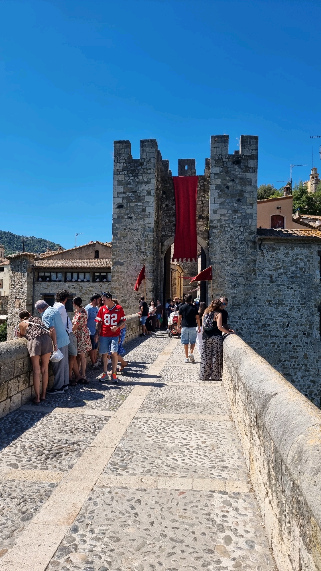 The image depicts the Pont Fortificat, a medieval fortified bridge in Besalú, Spain. The bridge is constructed of stone and features crenellated towers at its entrance, one of which prominently displays a large red banner. Cobblestones pave the bridge, with a lighter stone border running down the center.
A group of approximately 15-20 people are present on the bridge, some walking and others gathered along the parapet, appearing to observe something or take in the view. Among them, a man in a red shirt with the number "82" is a central figure. People are dressed in casual summer attire, indicating a warm day. The background shows stone buildings and the landscape typical of Besalú. The sky is clear and blue, suggesting fair weather. The overall scene captures a moment of tourism and exploration at a historical landmark.