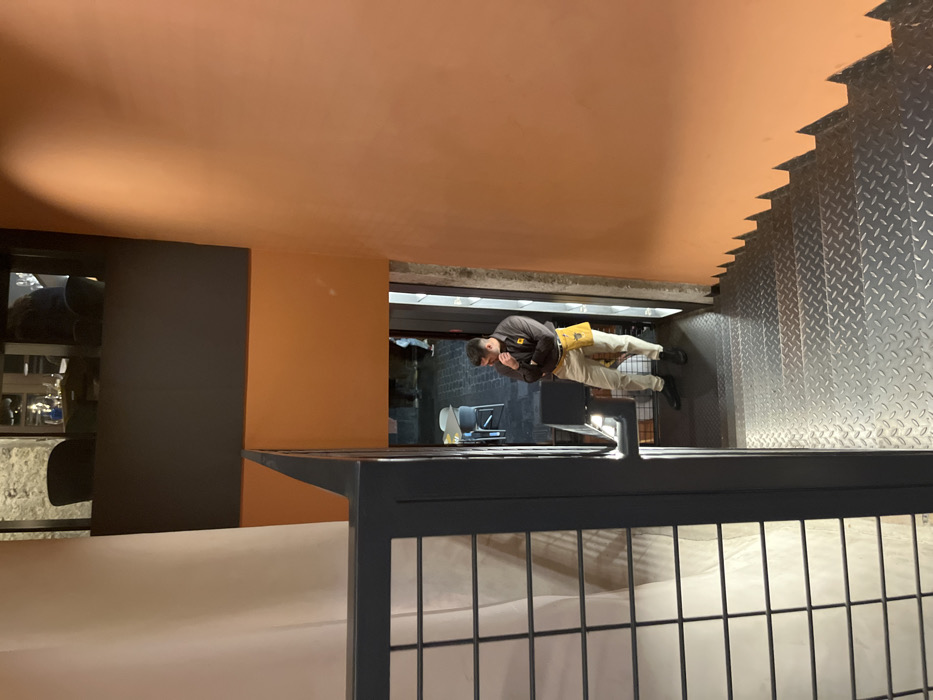 A man in a brown shirt and a yellow utility belt is standing at the top of a dark staircase. The man is looking down, his chin resting on his hand. His left leg is slightly bent, his right leg straight. The staircase leads down into a dimly lit space with a tiled floor. The walls are painted in various shades of orange. There is a black railing on the left side of the stairs. The image was captured in a building located in Las Palmas de Gran Canaria, Spain, at coordinates 28.102 latitude, -15.415 longitude. The scene is likely from a restaurant or a bar. The light comes from the top of the stairs.