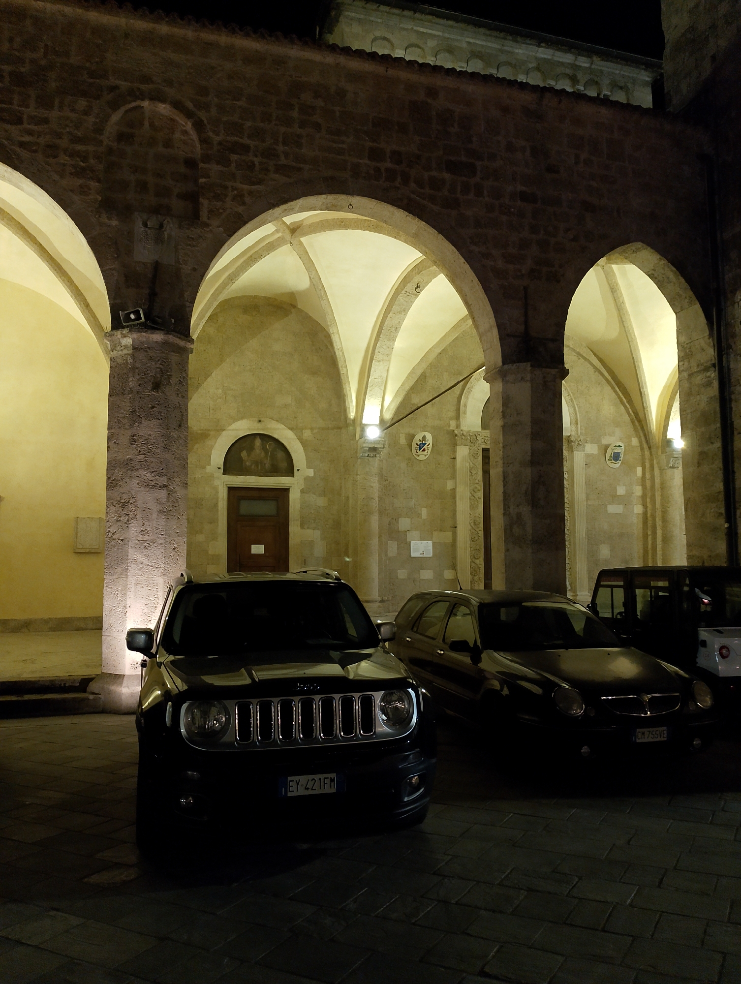 The image depicts an outdoor scene at night, likely in front of a historical building with arched walkways and stone architecture. Several cars are parked on a cobblestone surface. In the foreground, a black Jeep is prominently displayed, its headlights illuminated. To its right, a dark-colored sedan is parked, with a partial view of its license plate. Further to the right, a dark-colored SUV or van is visible, partially obscuring a white vehicle behind it. The building features arched openings and illuminated interiors, suggesting it might be a church or a public building. The overall atmosphere is one of urban nighttime ambiance, with artificial lighting casting shadows and highlighting the architectural features. The location is identified as Rieti, Italy, a city known for its historical sites and ancient architecture.