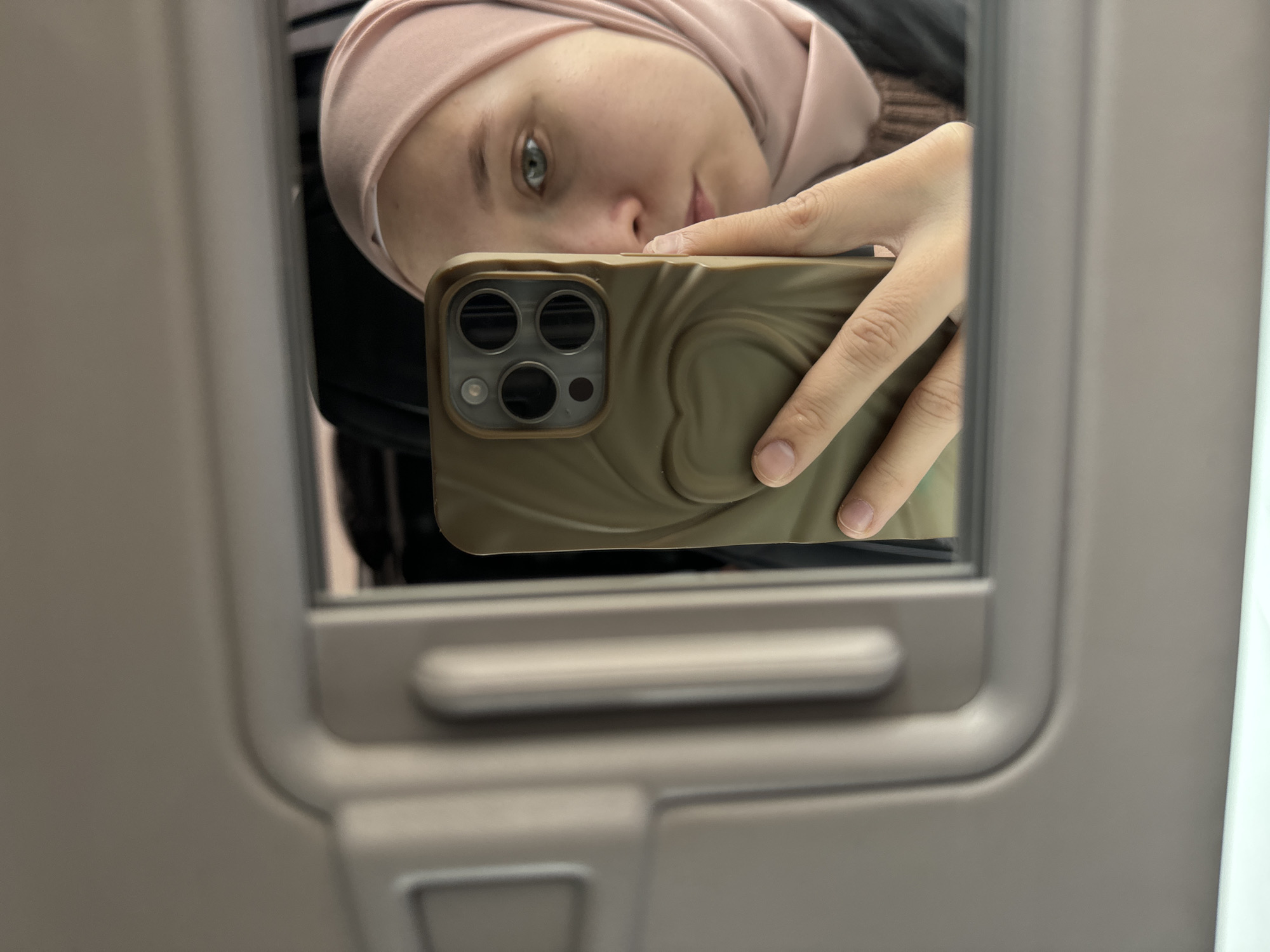 A person is taking a selfie in a mirror. The person is wearing a pink head covering, holding a smartphone with a brown case. The environment appears to be inside a vehicle, potentially a car or train, based on the interior paneling visible in the mirror's reflection. There is no information in the image to determine the location is Chaikovsky, Russia.