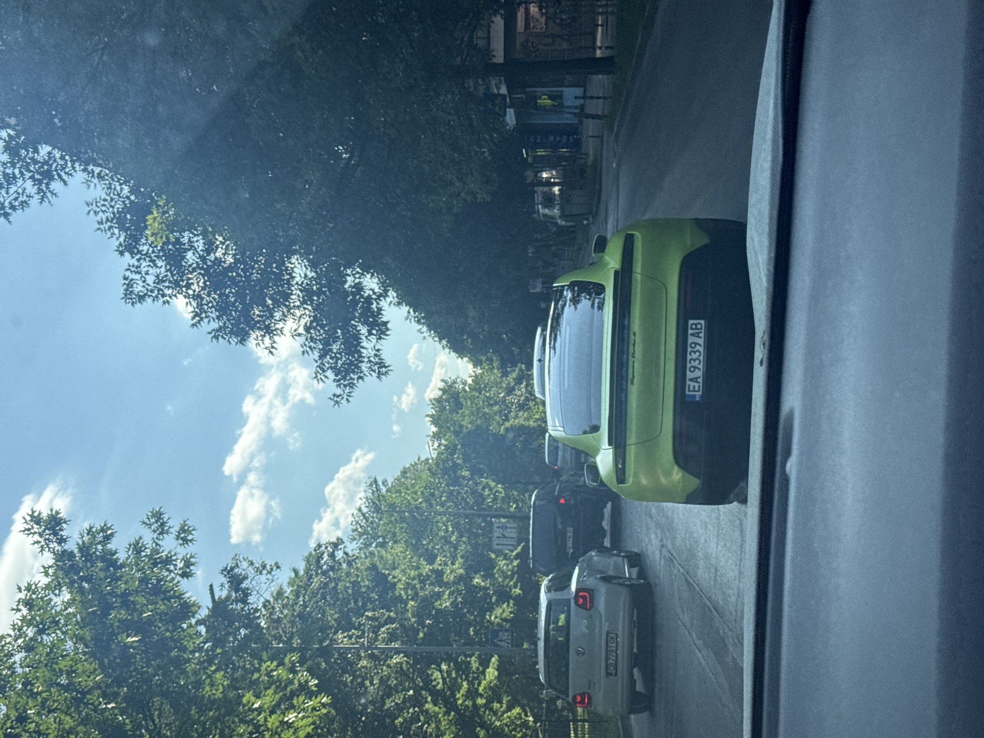 The image depicts a street scene in Sofia, Bulgaria, viewed from inside a vehicle. The road is lined with lush green trees on both sides, forming a canopy over the street. The sky above is blue with scattered white clouds, suggesting a clear day. Several cars are visible on the road, stuck in traffic. In the foreground, a bright green sports car is prominent, with its rear facing the camera. Its license plate reads "EA 9339 AB". To the left of the green car, a lighter colored car, possibly a Volkswagen Polo, is visible, with a license plate that appears to say "CB 7781 OK". Further down the road, other vehicles and a few shops with storefronts can be seen. The overall impression is a typical urban traffic situation on a sunny day in Sofia, with a focus on the vehicles and the surrounding greenery.