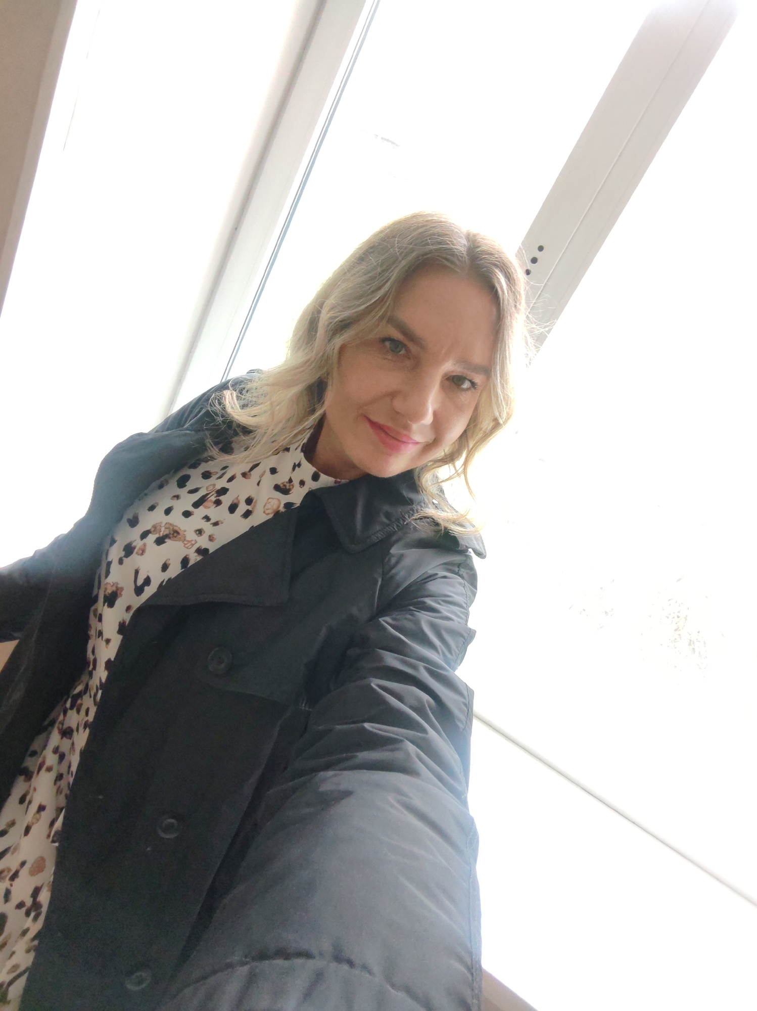 A woman poses indoors, wearing a light-colored dress with a dark-colored animal print and a black overcoat. The background consists of a bright, white wall with architectural features, suggesting a window or wall structure. The location is indicated as Unknown, Unknown.