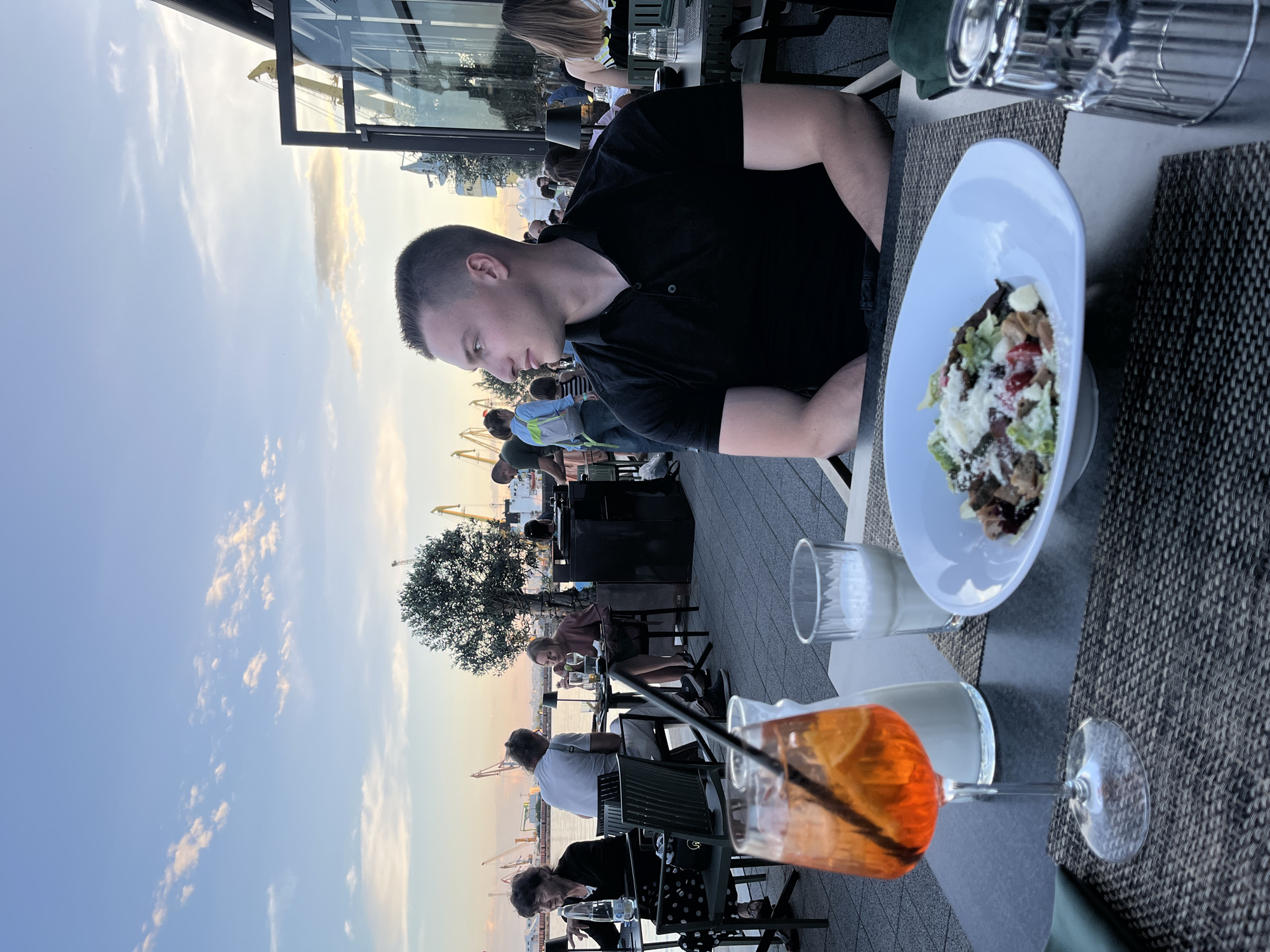 A man in a black polo shirt is seated at a table at an outdoor restaurant overlooking a port. In the foreground, a cocktail and a salad are on the table. In the background, several people are seated at other tables, and port cranes are visible against the sky. The scene is set at sunset in the port of Burgas, Bulgaria.