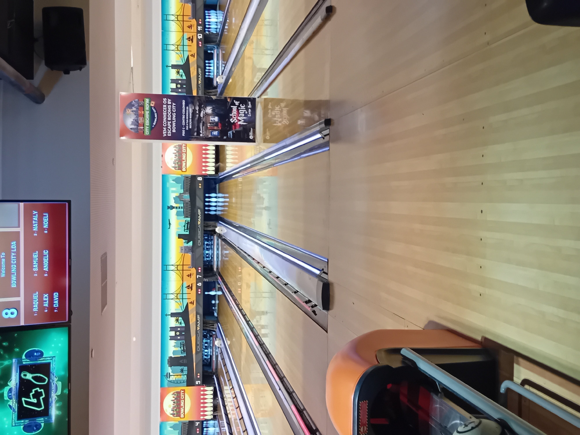 The image captures an indoor bowling alley, "Bowling City LDA," likely located in Lisbon, Portugal, given the Portuguese text on the "City Escape Room" poster. The lanes are numbered and equipped with automatic scoring systems. Advertisements for escape rooms and "School of Magic Escape Room" are displayed. A screen shows player names: RAQUEL, SAMUEL, NATALY, ALEX, ANGELIC, DAVID and NOELI. An electronic display indicates a score of "48".