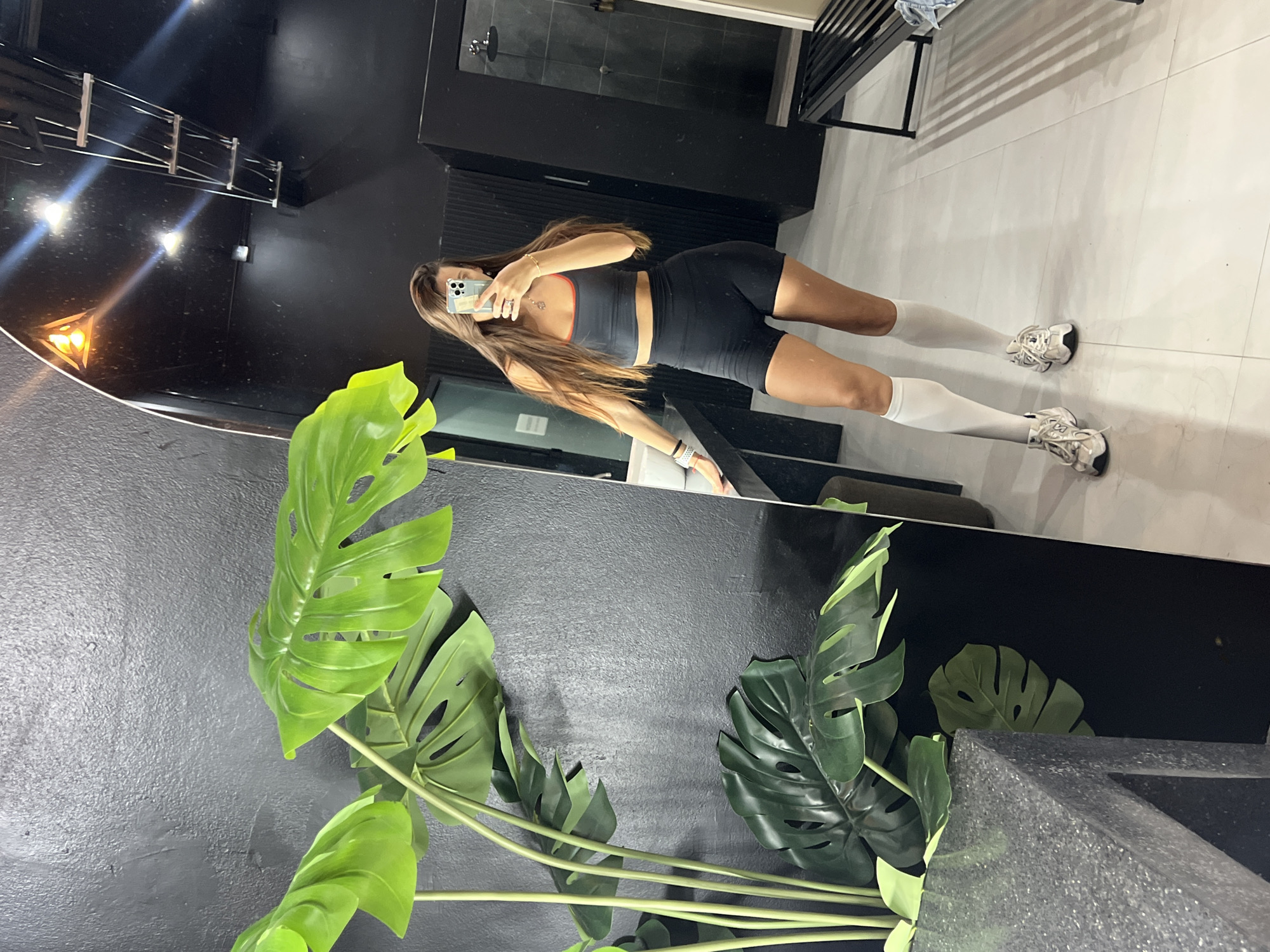 A mirrored reflection captures a woman in workout attire holding a phone up to her face, seemingly taking a photo of herself. She is wearing a dark sports bra, black shorts, and white socks with athletic shoes. The reflection shows a minimalist bathroom with black walls and light-colored tile flooring. A metal rack is visible on the right side of the reflection. In the foreground, there is a large plant with green leaves, adding a touch of nature to the scene. The image is rotated, with the plant and mirror positioned on the left and the hallway with the woman on the right.