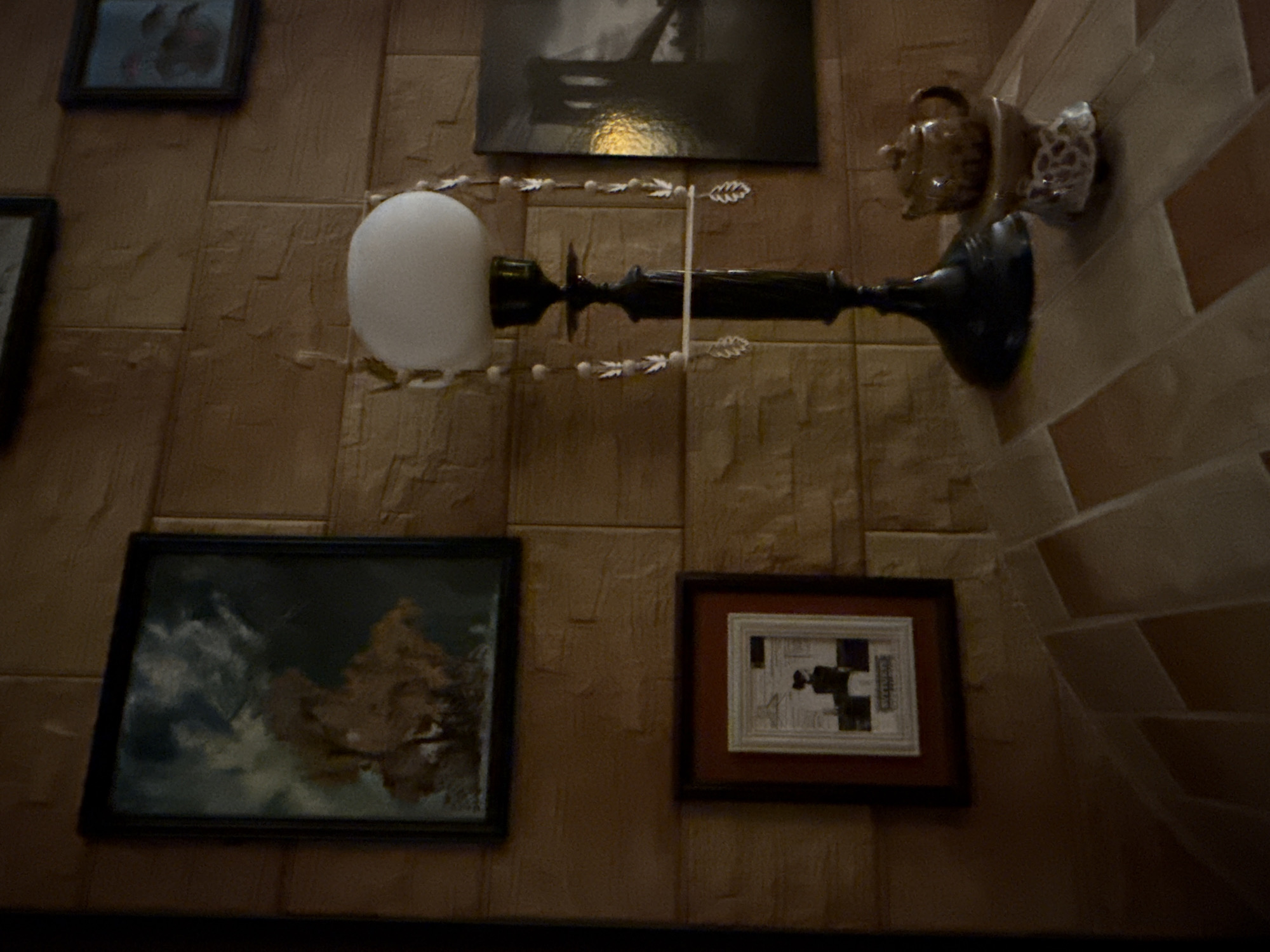 The image depicts an indoor scene, likely a residential interior, with a brick-patterned wall as the backdrop. On the wall, there are three framed pictures. The largest and leftmost picture is a painting with abstract brushstrokes in shades of blue, brown, and white, possibly depicting a landscape or a stormy sky. To its right, a smaller framed picture contains a black silhouette of two figures against a light background, with some text below. The third framed picture, on the far right, is a black and white photograph of a bridge, with a yellow light source reflected on its surface.
In the foreground, on a surface with tiled sections in shades of brown and beige, there is a black lamp with a white globe shade. Decorative beads with leaf-shaped pendants hang from the lamp. Next to the lamp, a small brown teapot sits on a decorative ceramic stand with an intricate pattern. The overall ambiance is dimly lit and somewhat vintage.
The image provided does not contain any information or visual cues that would allow for the identification of its location within the city of Galinė, Lithuania.