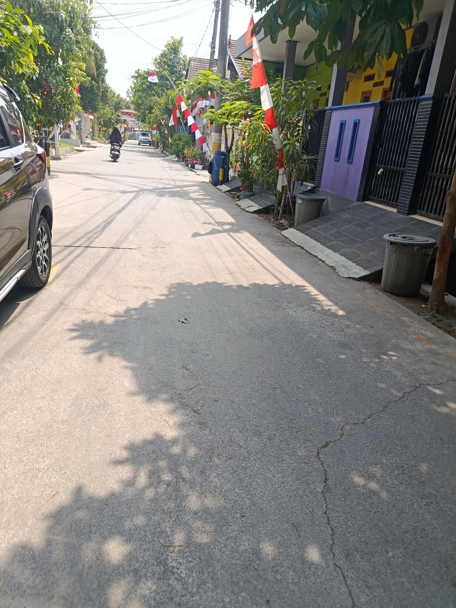 The image depicts a street scene in Wanasari, Indonesia. A dark-colored SUV is parked on the left side of the road, partially obscuring a portion of the street. A person on a scooter, wearing a black hijab and dark clothing, is riding away from the camera down the street. The street is lined with houses on the right side, many of which are decorated with Indonesian flags. Trees and plants are visible along the sidewalks and in front of the houses. The road surface is asphalt and shows signs of wear, with visible cracks. Shadows from trees are cast across the road, indicating a sunny day. Two grey trash bins are placed on the right side of the road near a ramp leading to a house. The sky is clear and bright.