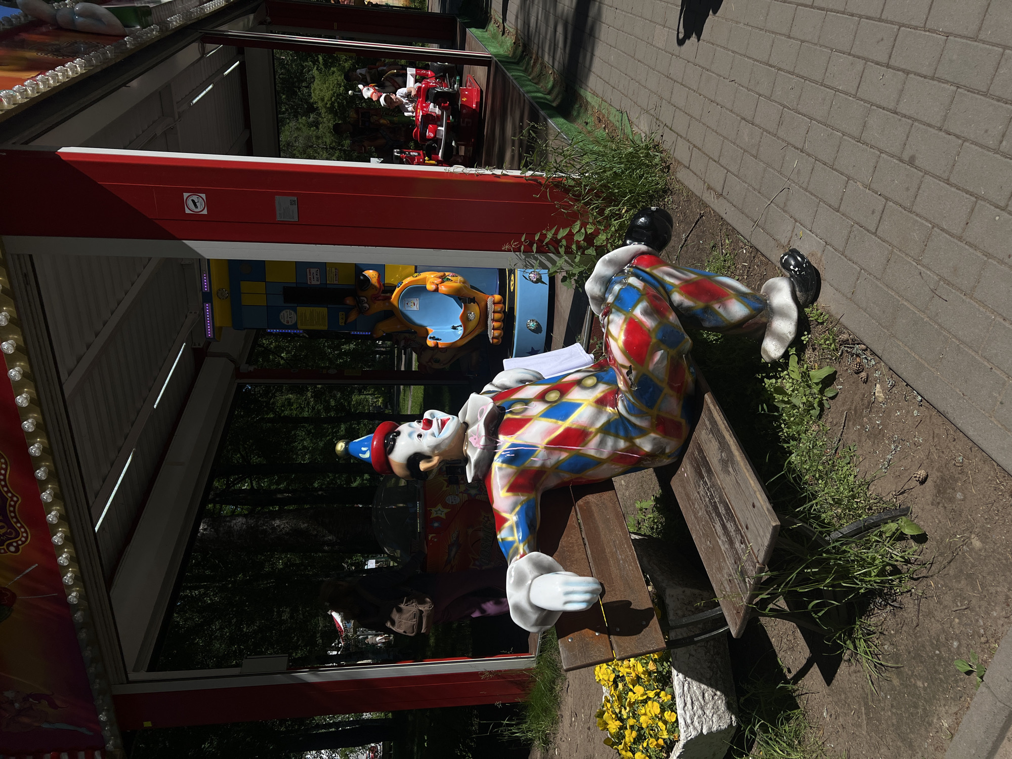 The image depicts an outdoor amusement park scene in Zelenogorsk, Russia. In the foreground, a life-sized clown statue is seated on a wooden bench. The clown wears a colorful diamond-patterned costume in red, blue, and white, a red and blue cone hat, and has a painted white face with a red nose and smile. The bench is weathered and surrounded by sparse green grass and a planter filled with yellow flowers. Behind the clown, a red and white amusement ride structure is visible, with children on the ride in the background. The ground is a mixture of dirt and a paved walkway made of grey rectangular bricks. The scene is illuminated by sunlight, casting shadows.