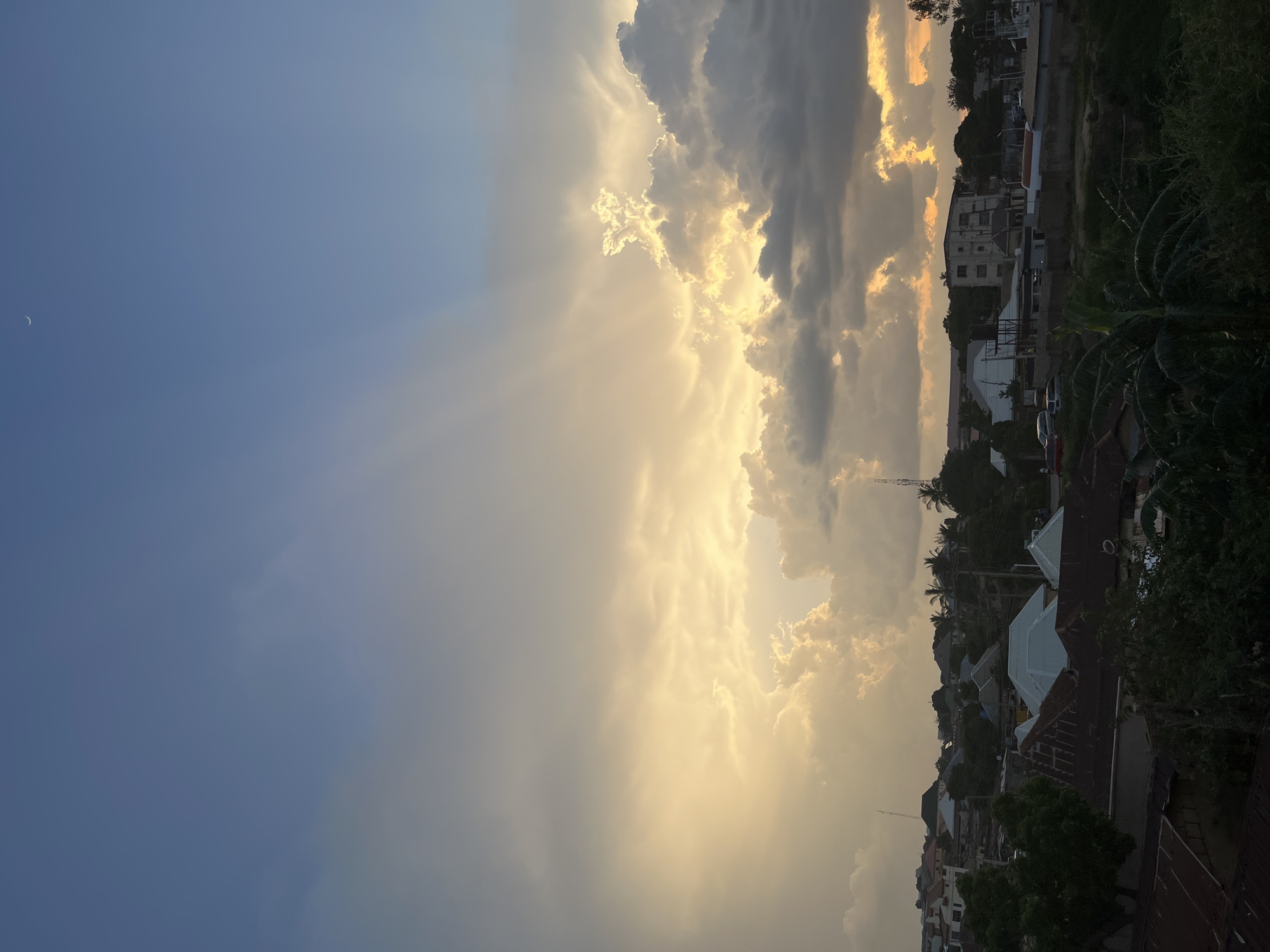 This media file shows a landscape view of Jos, Nigeria, during what appears to be sunset. The sky is dramatically filled with a mix of dark and bright clouds, with the sun's rays piercing through, creating a dramatic effect. On the left side of the frame, a faint crescent moon is visible in the clear blue sky. The right side of the image shows a town with houses and buildings, some with distinctive red or white roofs. Dense vegetation, including palm trees, is visible throughout the scene. The lighting suggests a warm, possibly humid, evening. The overall mood is serene and picturesque, capturing the beauty of the African sky at dusk. There is no visible text in the image.