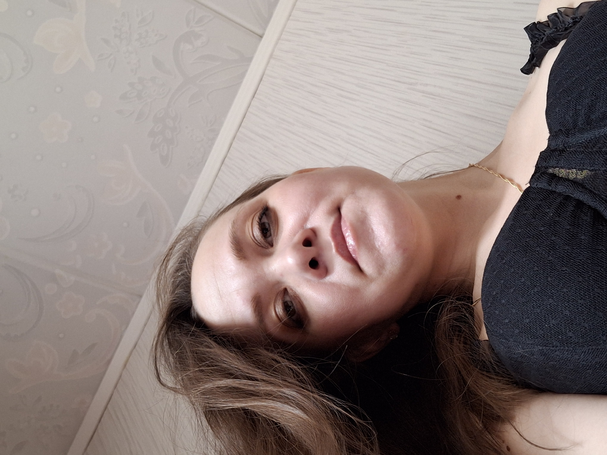 A woman with brown hair and fair skin is seen from a low angle, her face close to the camera. She has brown eyes and glossy lips. She is wearing a black top with a textured pattern and a small ruffled detail on the shoulder. A delicate gold necklace is visible around her neck. The background shows a wall with light-colored wood paneling and a patterned wallpaper. The image was most likely not taken in the city of Chaikovsky, Russia, as this detail is not discernible in the image.