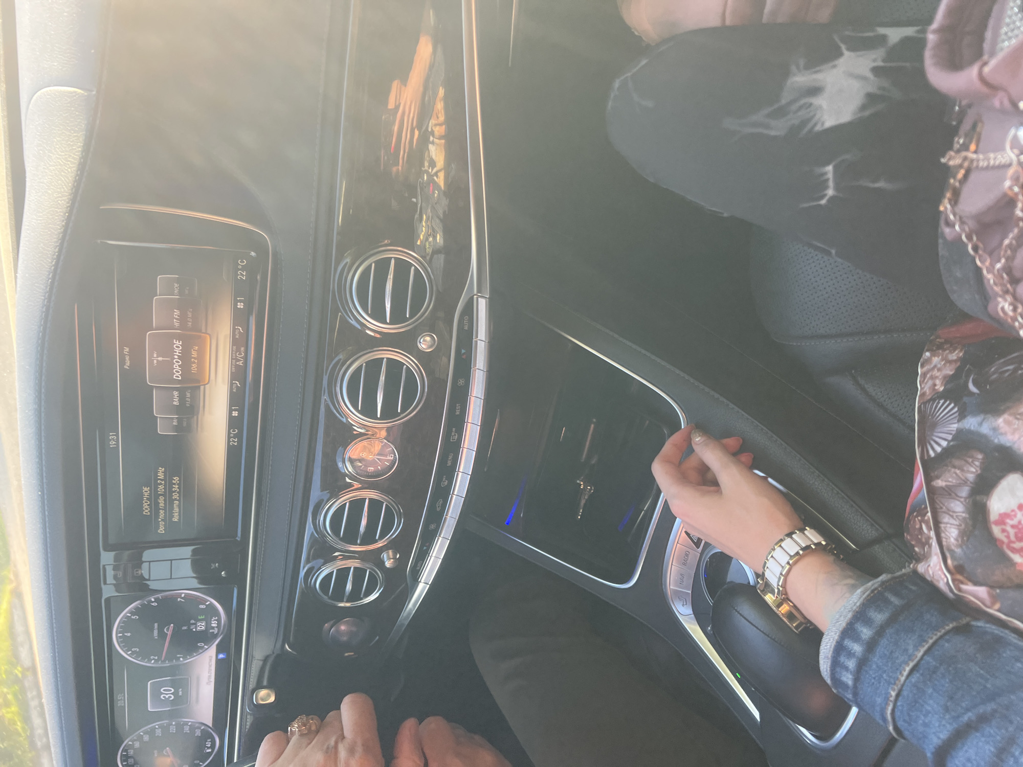 This is a candid, eye-level shot taken from inside a car, focusing on the dashboard and the occupants. The time is 19:31, and the weather appears to be warm with a visible temperature of 22°C on the car's display. The scene is set in Krasnoe-na-Volge, Russia, as indicated by the text on the screen.
The primary subjects are two individuals, seen from the torso down. One person, whose hands are visible in the foreground, wears a large, ornate ring and is placing their hand near the gear shift. The other individual, in the background, is wearing a denim jacket and a watch with a white and gold band. Their jeans have a distinctive ripped pattern. A patterned handbag with a gold chain rests on the passenger seat.
The car's interior is well-lit, with the dashboard featuring a prominent infotainment screen displaying radio station information, including "DOPO*HOE" on 106.2 MHz and "HIT FM." Other visible text includes "Радио FM," "Reklama 30-34-56," "AUTO," "A/C," and a speed reading of "30." The car's climate control also shows "22°C." The overall mood seems relaxed, suggesting a casual drive or journey. The bright sunlight filtering into the car hints at either late afternoon or early evening, depending on the season.