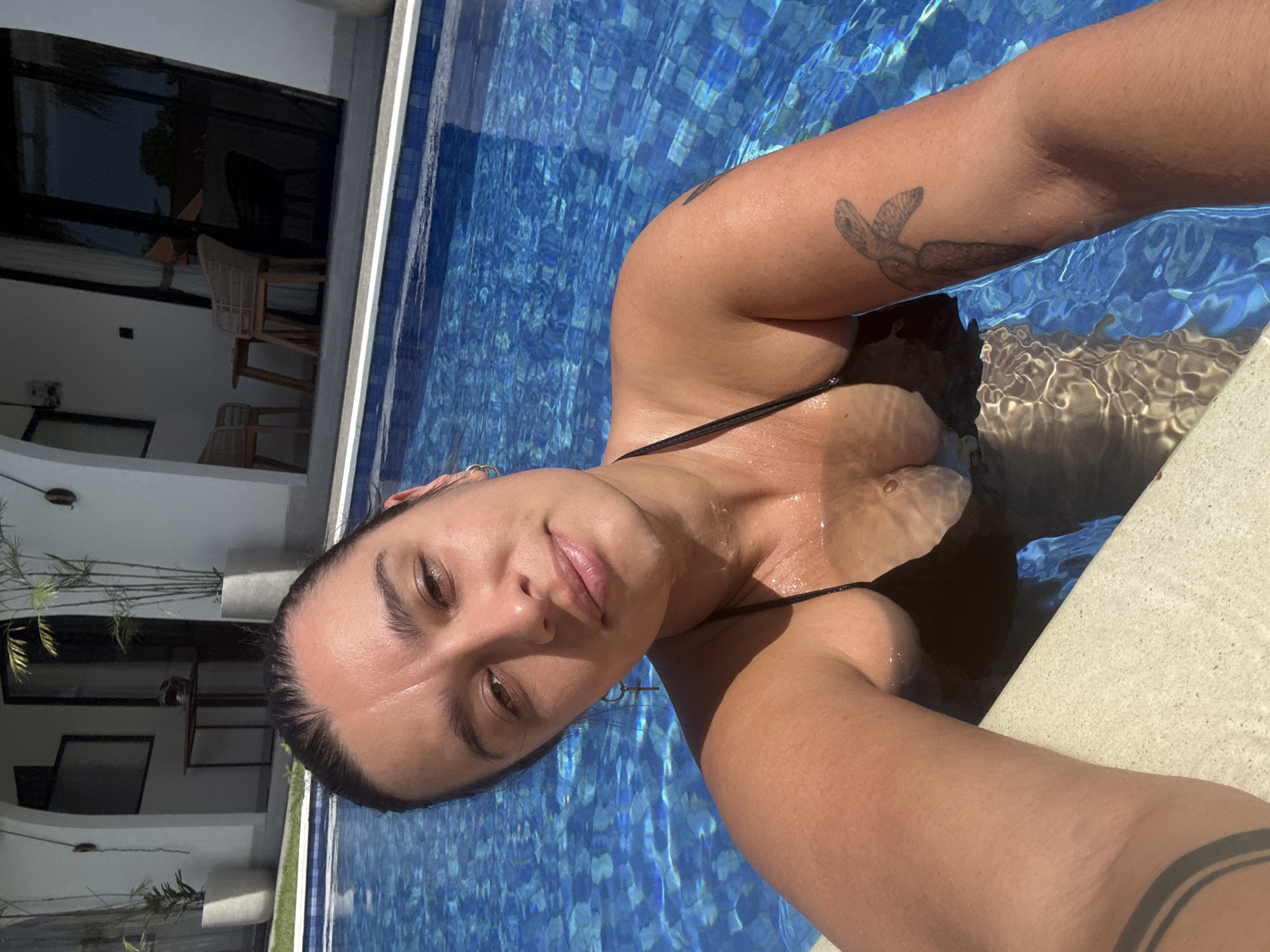 The image depicts a young adult female taking a selfie while submerged chest-deep in an outdoor swimming pool at a location in North Kuta, Indonesia. The female has dark hair pulled back, a tanned complexion, and is looking directly at the camera with a slight smile. She wears a black bikini top and has a stylized dark-colored turtle tattoo on her right upper arm and a dark band tattoo on her right wrist. A small hoop earring with a cross charm hangs from her left ear, and a simple hoop is in her right ear. Her forearms rest on the light-colored coping of the pool. The pool water is clear, revealing blue tiles at the bottom, and reflects bright sunlight.
In the background, behind a narrow strip of green grass, is a white-walled building with resort-style architecture, featuring arched doorways and dark-framed glass doors or windows. On a patio area in front of the building, two woven chairs and a small table are visible. Tall, potted tropical plants flank an entryway. The entire scene is brightly illuminated by direct sunlight, indicating clear weather.