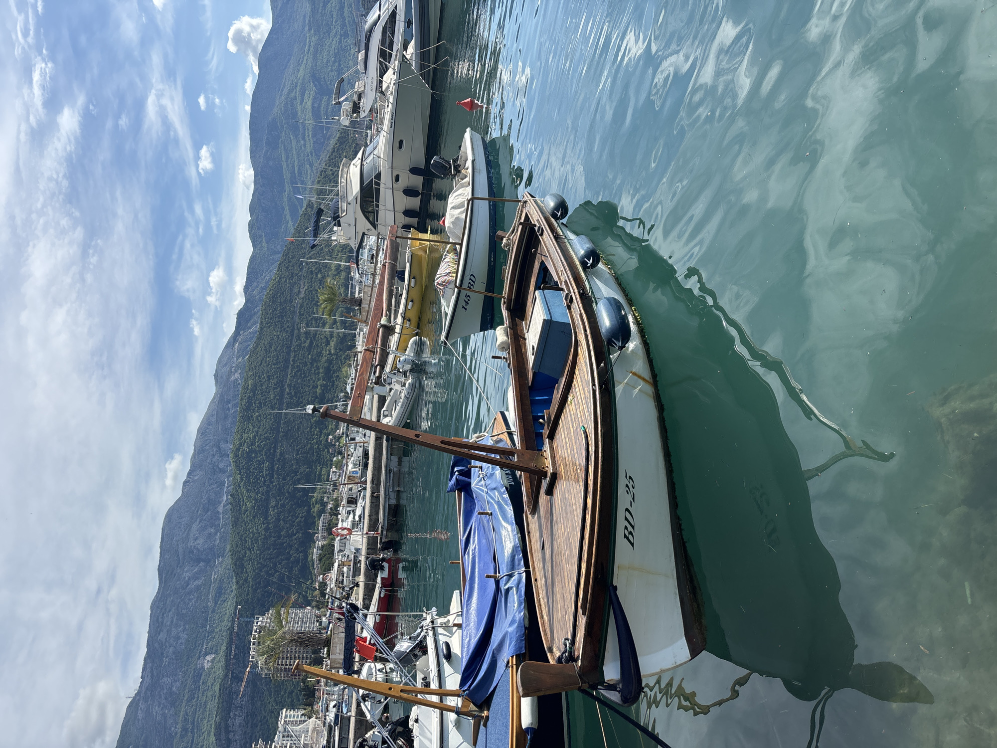 The image shows a marina in Budva, Montenegro, with several boats docked. In the foreground, there is a small wooden boat with a blue cover, bearing the registration number BD-25. Behind it, there are other boats, including a larger yacht. The water is calm and reflects the boats and the sky. In the background, there are mountains and some buildings of Budva. The sky is partly cloudy. The picture appears to be taken during daytime with good lighting.