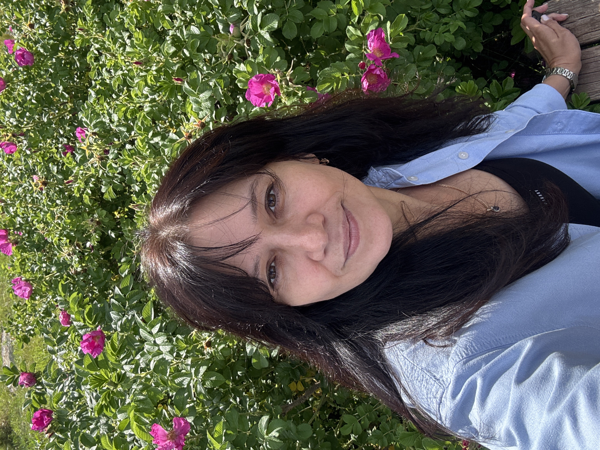 A woman with long, dark hair and brown eyes smiles at the camera, posing for a selfie in front of a blooming rose bush. She is wearing a light blue button-up shirt and a black tank top. Her right hand rests on a wooden surface, possibly a bench. The background is filled with vibrant green leaves and bright pink flowers of a rose bush. The scene appears to be outdoors, likely in a park or garden, and the lighting suggests it is a sunny day. The image has been cropped to focus on the woman and the flowers. Without further information, it is not possible to determine the exact location in Chaikovsky, Russia.