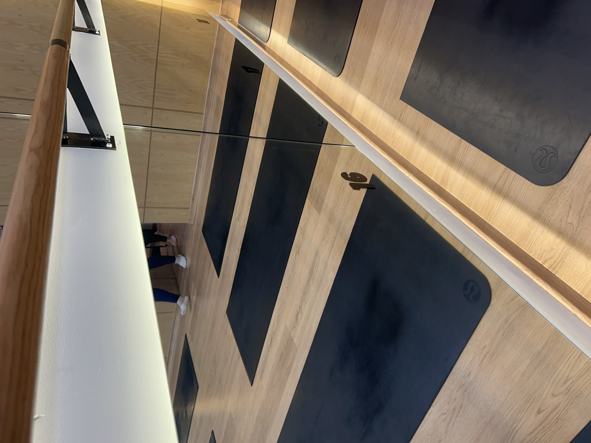 The vertical image depicts an interior scene, likely a fitness studio, dominated by reflections in a large mirror. In the foreground, along the left side of the image, is a light-colored wooden barre attached to a white, vertical partition or wall structure by black metal brackets.
The majority of the frame is filled by the large mirror, which reflects the room beyond. Reflected in this mirror is a light wooden floor where several black exercise mats are neatly arranged in rows. Each mat clearly displays a circular logo, identifiable as the Lululemon emblem. The reflection also shows the lower halves of people; specifically, a pair of legs wearing blue pants and white athletic shoes or socks is visible, indicating someone is standing on or near the mats.
The presence of the barre and the exercise mats suggests the setting is a studio used for activities such as barre, yoga, or Pilates. The lighting is bright and even, typical of an indoor facility. No specific time of day, weather, or discernible emotions are apparent. The primary visible text or branding is the Lululemon logo on the mats.