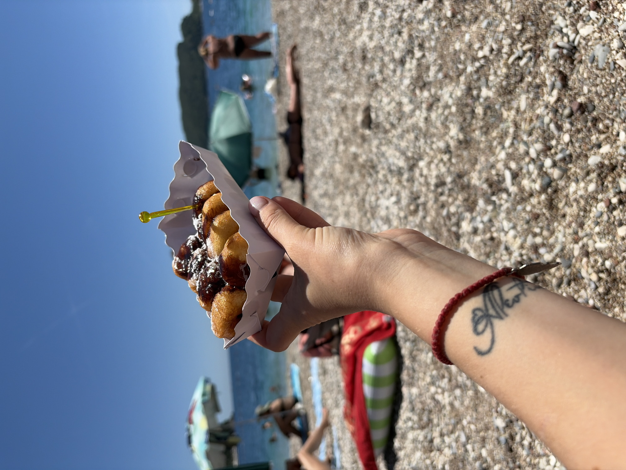 In Budva, Montenegro, a hand holds a white paper container filled with donut holes dusted with powdered sugar and drizzled with chocolate sauce, topped with a yellow plastic toothpick. The scene unfolds on a pebbled beach under a clear blue sky. In the background, the turquoise Adriatic Sea stretches out, with a distant headland visible. Beachgoers relax on towels and swim, some under colorful umbrellas. One person stands near the water, facing away from the camera. The foreground shows the pebbled beach and the tattooed forearm of the person holding the donuts. A thin red bracelet is worn on the wrist.