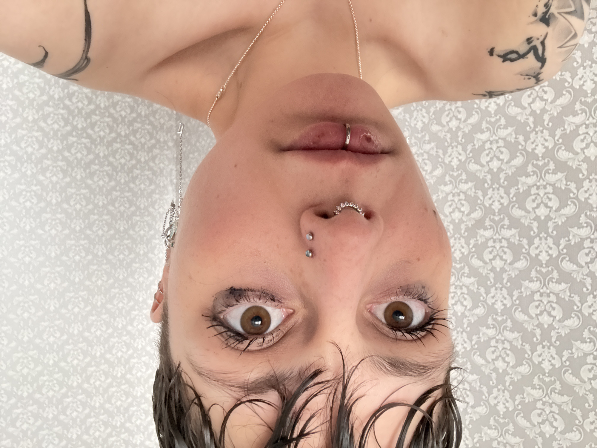 A person is photographed upside down against a backdrop of grey and white patterned wallpaper. The person has fair skin, short dark hair partially obscuring the forehead, and brown eyes that are looking upwards toward the camera. They have multiple piercings, including a ring in their lower lip, a jeweled ring in their nose, and two small studs between their eyebrows. A silver chain necklace is visible around their neck, and decorative earrings are visible on one ear. The upper arms and shoulders are visible, and tattoos are present on both arms. The photo does not contain any information about the location being in Saint Petersburg, Russia.