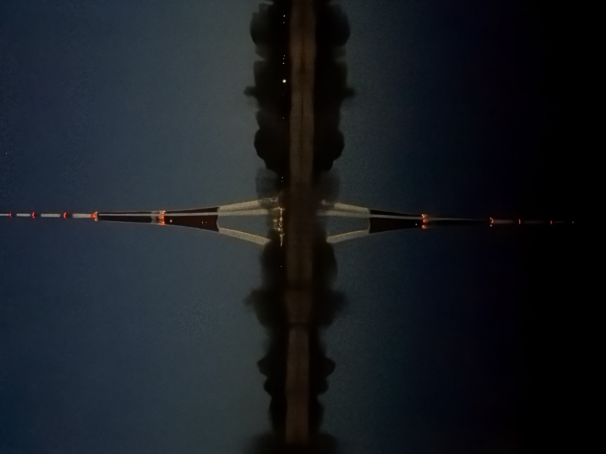 The image depicts a symmetrical composition of what appears to be the reflection of a bridge and its surroundings in water, possibly at night. The bridge's structure, possibly a cable-stayed bridge, stretches horizontally across the frame, reflected in the water below. The central vertical axis features a blurry, dark shape that likely represents the bridge's reflection in the water, creating a somewhat distorted image. Tiny bright lights are visible in this vertical shape. The bridge structure itself appears to have lights along its span, some appearing red. The background is a dark hue, possibly the night sky. Based on the bridge structure and the lighting, it could be the Vanšu Bridge in Riga, Latvia.