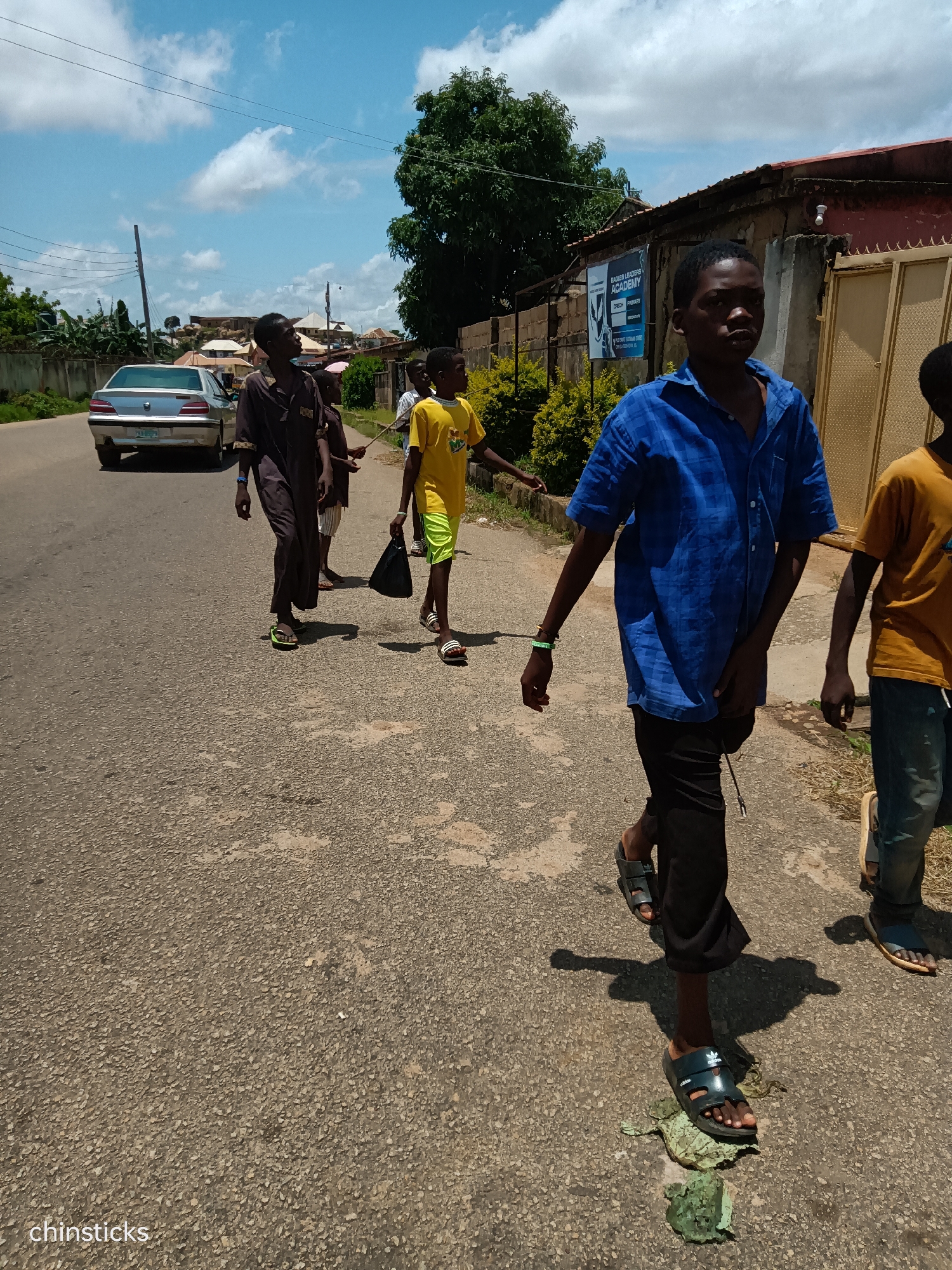 In Jos, Nigeria, on a paved road under a bright, cloudy sky, a group of boys walks along the side of the road. The boy in the foreground, wearing a blue checkered shirt and black pants, is in motion, his shadow stretching across the asphalt. He wears black sandals, one of which is positioned on a pile of green debris. To his right, another boy in a yellow t-shirt and blue jeans walks, his shadow also visible. Further behind, a boy in a dark brown robe and another in a yellow t-shirt with lime green shorts are walking together, the latter carrying a black bag. A silver sedan is parked on the left side of the road, its rear facing the camera. A building with a sign that reads "EAGLES LEADERS ACADEMY" is visible in the background, alongside a tall tree and other structures. The environment appears to be a moderately urban or semi-urban area.