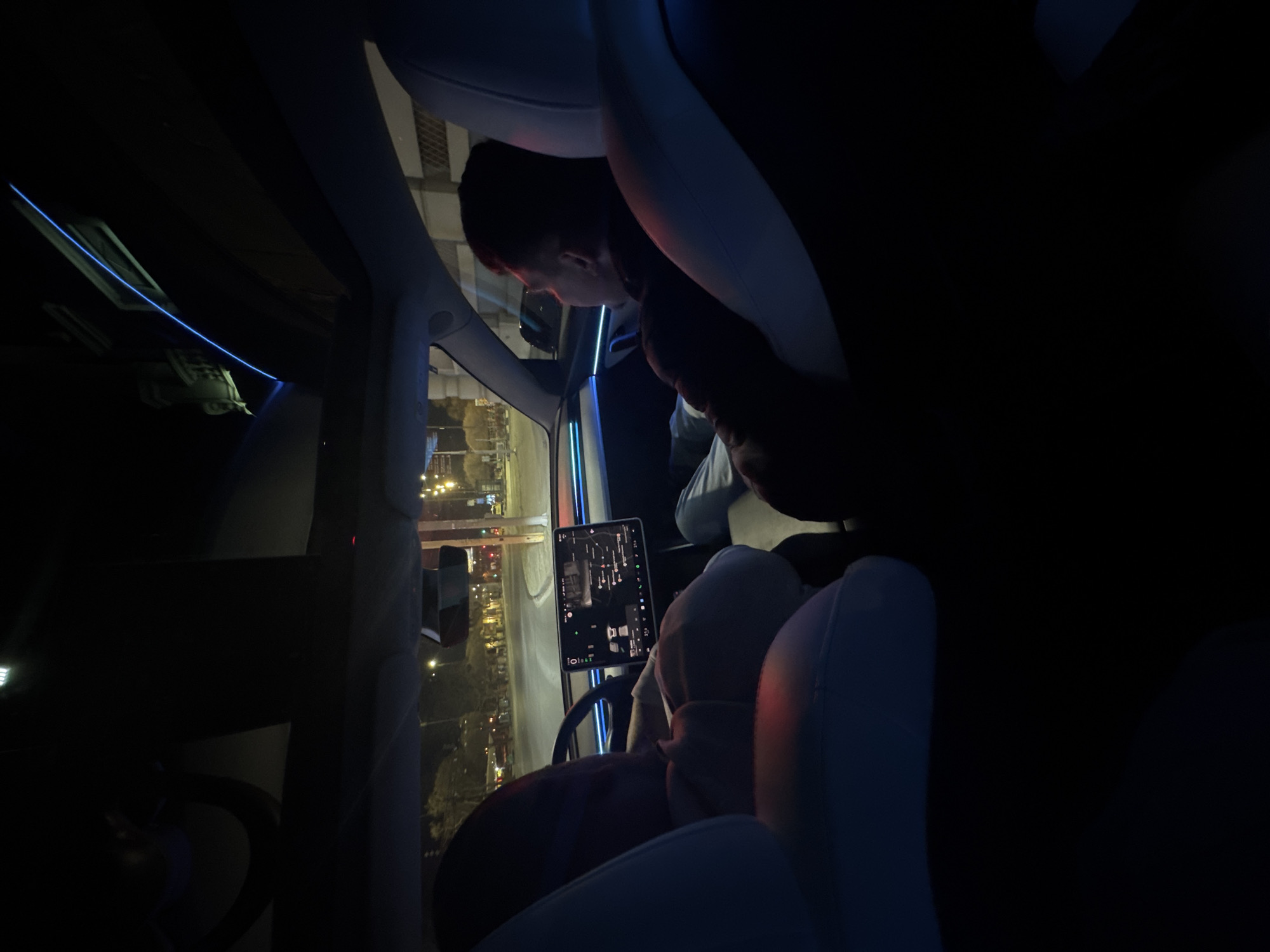 The image depicts the interior of a car, likely a Tesla, with the driver looking out the window. The car appears to be parked or stopped, as the screen is illuminated with a map. The location is identified as Varna, Bulgaria, based on the streetlights and buildings visible through the car window. The interior of the car is illuminated with ambient blue light. The person in the car has short, dark hair and is wearing a dark shirt and white or grey long sleeved shirt. The seats are white and appear to be leather or a similar material. The image is taken from the back seat of the car, pointing towards the driver and the road outside.