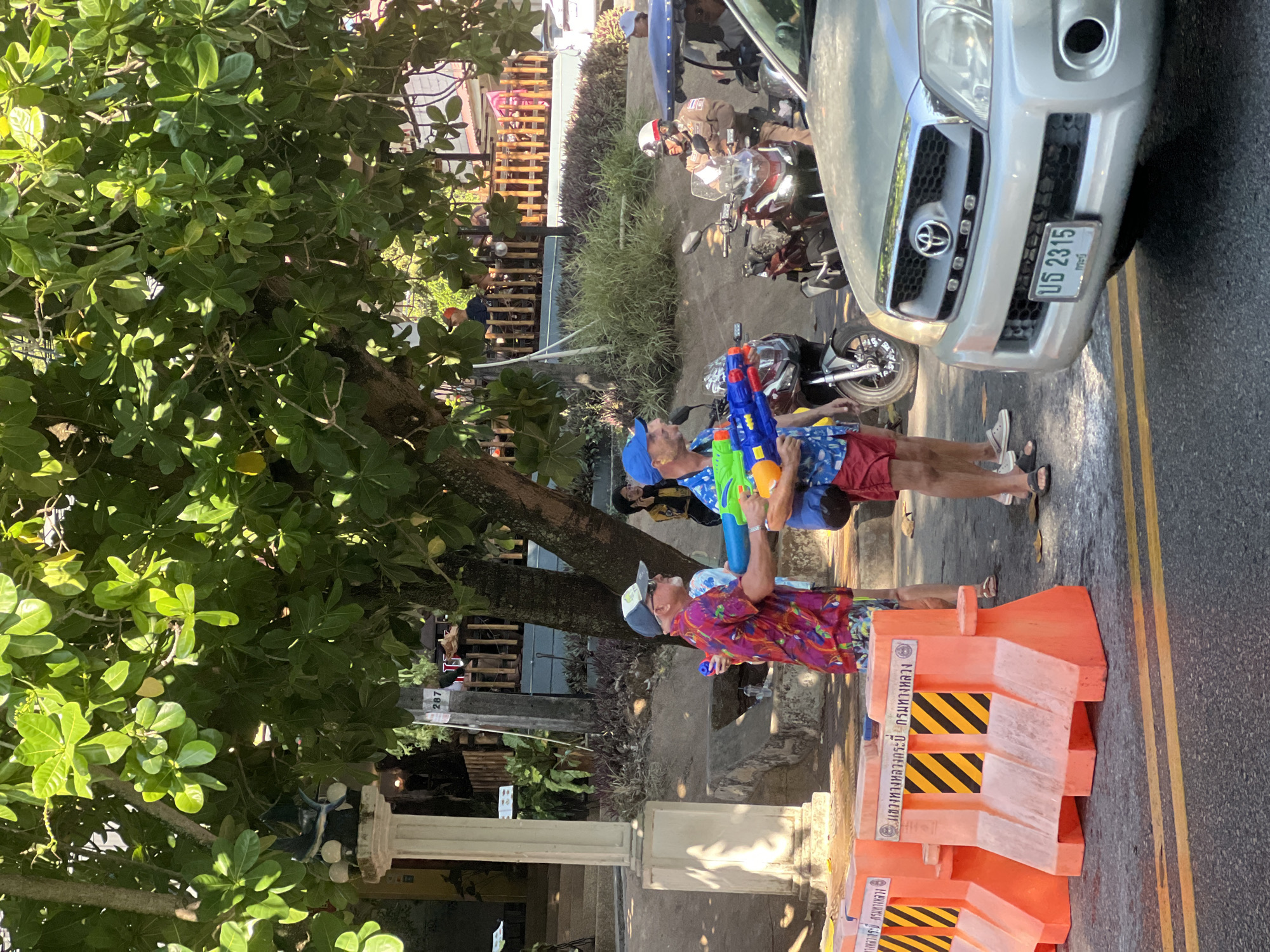 This image depicts a street scene in Ban Klong Haeng, Thailand, featuring two men participating in Songkran. They are positioned near orange and black safety barriers along the road. Both men are holding water guns, suggesting they are engaged in water play, a common activity during Songkran. The man closer to the camera wears a colorful shirt and a hat, while the other man wears a blue hat. Parked nearby are several motorcycles and a silver Toyota pickup truck, with the license plate บธ 2315 visible. A large tree with dense foliage is located on the left, and a building with a marker post (287) is visible in the background.