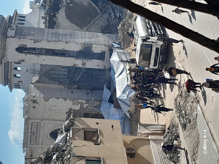 The image depicts a scene of extensive damage and humanitarian activity in Khan Yunis, Palestinian Territory. In the background, a large, multi-story building, identified by its dome and arched windows and entrance, appears significantly damaged, with large sections of its facade missing and internal structures exposed. Adjacent to it, a tall, slender structure, possibly a minaret, is also heavily damaged and partially collapsed.
In the mid-ground, a cluster of makeshift shelters constructed from white tarpaulins is visible on a dirt patch. A substantial crowd of people, including adults and children, is gathered around these tents and near a white Volvo tanker truck with a metallic cylindrical tank, suggesting the distribution or collection of water or other supplies. Many individuals in the crowd are holding buckets and containers.
The foreground shows a paved street bordered by piles of rubble and debris from destroyed structures. Several individuals are walking on this street; one person is bent over near a rubble pile. To the left, a multi-story building with an air conditioning unit mounted on its exterior shows signs of damage, with its upper section appearing to be in disrepair or partially collapsed. The sky is partly cloudy, and the scene is illuminated by natural light, casting shadows.