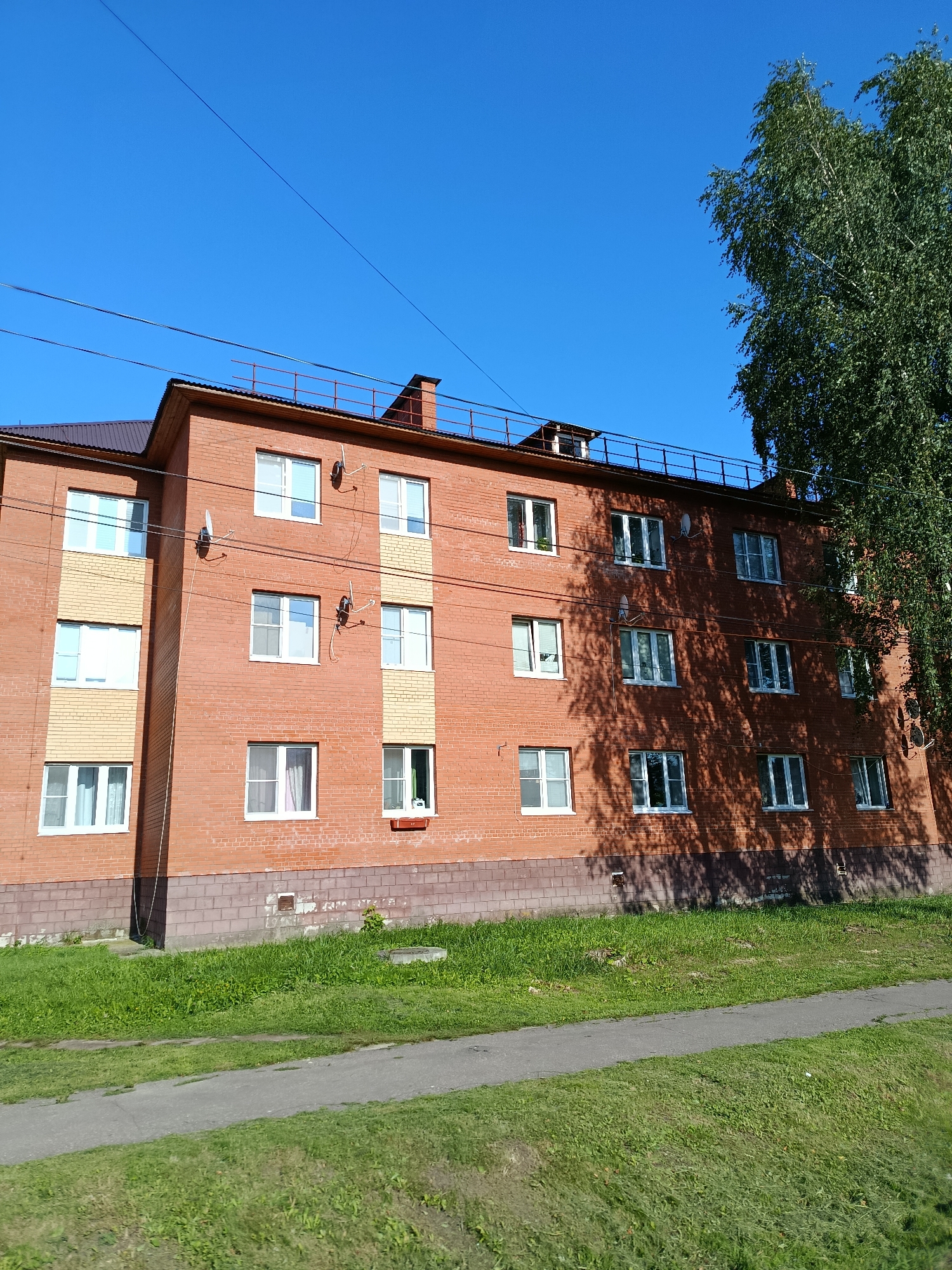 The image depicts a multi-story brick apartment building located in the city of Vereya, Russia. The building has a brown roof and numerous windows, many with white frames. There is a sidewalk and a grassy area in front of the building, with a large tree on the right side partially obscuring the structure. Power lines are visible in the sky above the building. The scene appears to be under a clear blue sky.