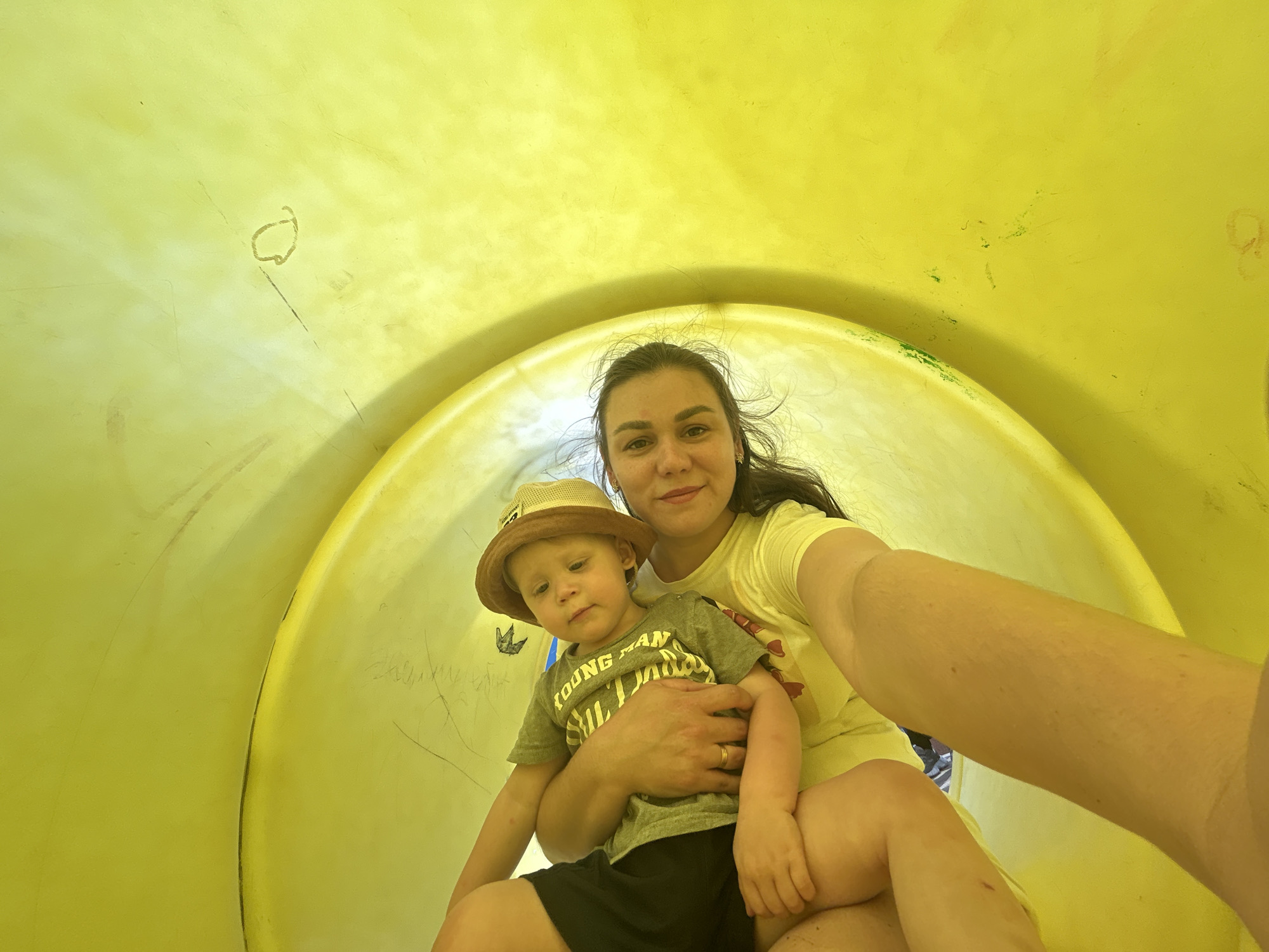 A low-angle, wide shot captures a woman and a young boy sitting in a bright yellow, enclosed slide. The woman, in a yellow t-shirt, holds the boy, who is wearing a tan bucket hat and a grey t-shirt with the words "YOUNG MAN" visible, on her lap. They are positioned within the curved interior of the slide, suggesting they are either at the start or end of a descent. The background is entirely the smooth, plastic surface of the slide, with some faint scratches and smudges visible. The lighting is bright and even, indicative of daylight. The location is identified as the city of Chaikovsky, Russia.