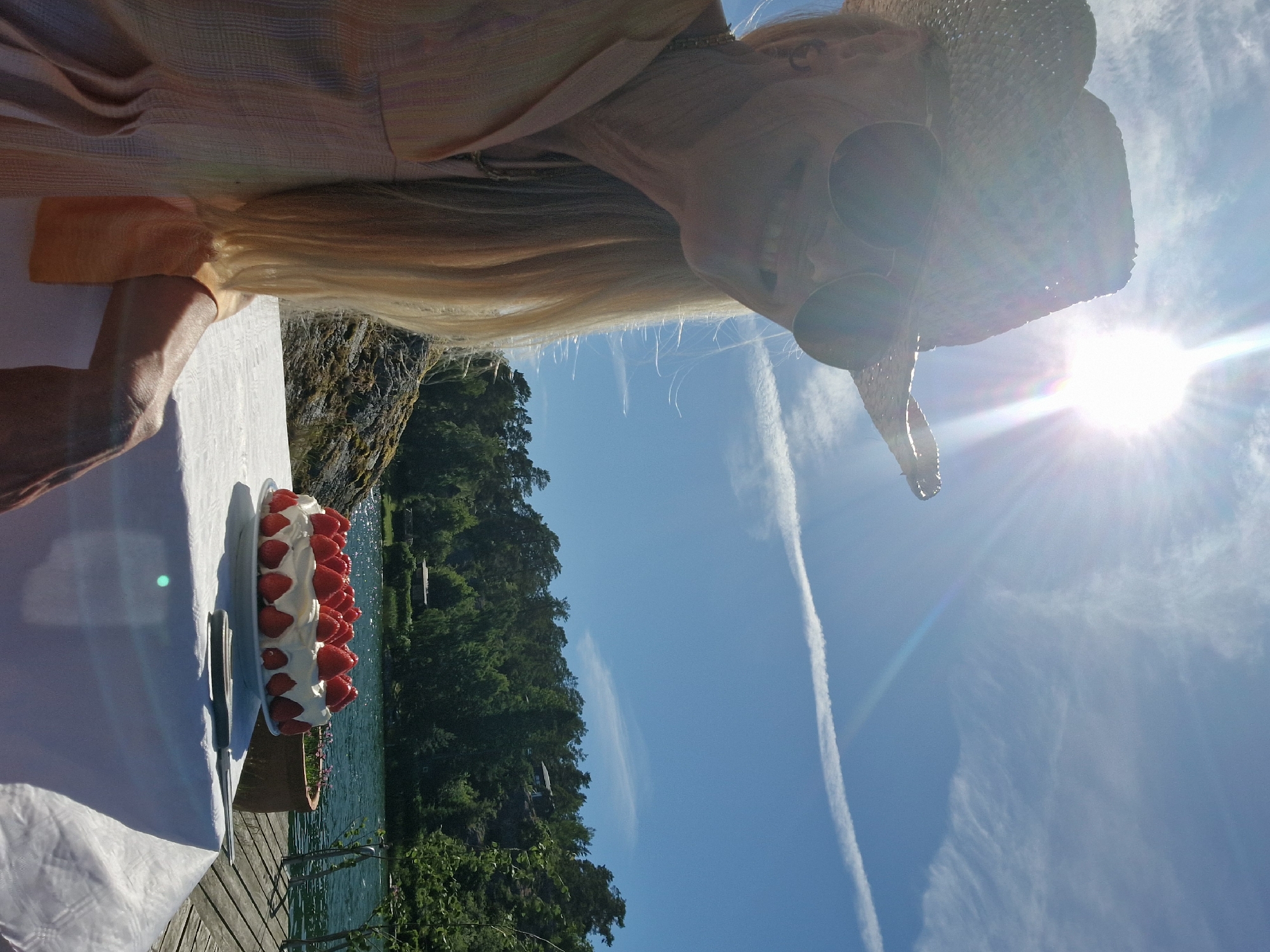 This is a medium shot of a person in a straw hat and sunglasses, holding a strawberry cake. The person is wearing a light-colored top. The cake is round and covered in whipped cream and fresh strawberries. It sits on a white tablecloth on a wooden table. To the left of the cake is a knife. In the background, there is a body of water, likely a lake or sea, with green trees on the opposite shore. The sky is bright blue with some wispy clouds and a sun flare. The scene appears to be outdoors, possibly on a deck or pier, given the wooden planks visible. The time of day is likely midday, judging by the bright sun and shadows. The overall mood seems festive and relaxed, suggesting a celebration. The image is oriented vertically, with the person's head near the top and the cake towards the bottom.