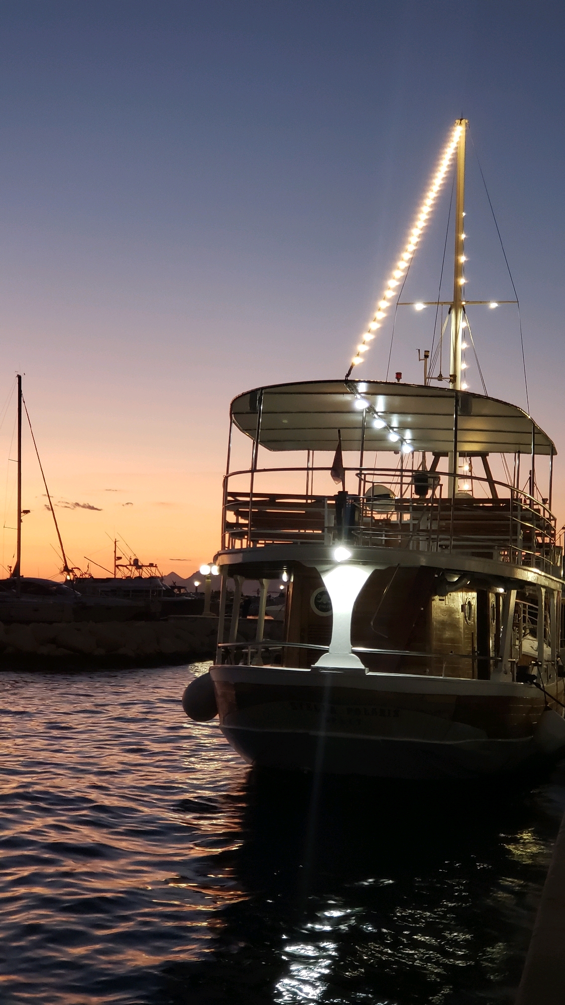 A wooden boat with multiple levels is moored in the harbor of Općina Tučepi, Croatia, at dusk. The boat is illuminated with strings of lights along its mast and deck railings. The water in the foreground reflects the lights and the warm hues of the sunset. In the background, other boats and a silhouetted coastline are visible. The sky transitions from a pale orange at the horizon to a deep blue overhead. There are no people visible in the image.
