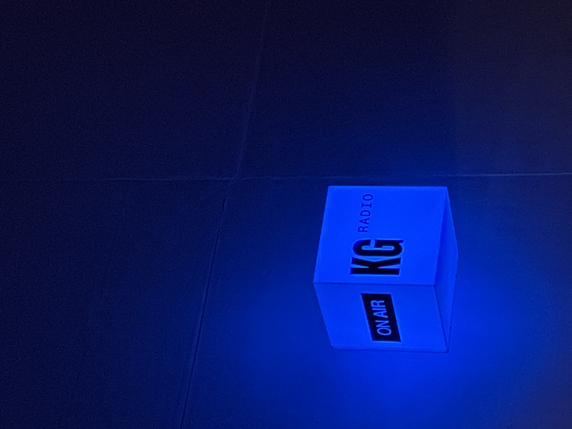 A top-down view of a lit blue cube, likely an "on air" light, on a dark tiled floor. The cube is labeled "KG Radio" and "On Air" in black lettering. The location, Belgrade, Serbia is not discernible from the image. The environment is dark.