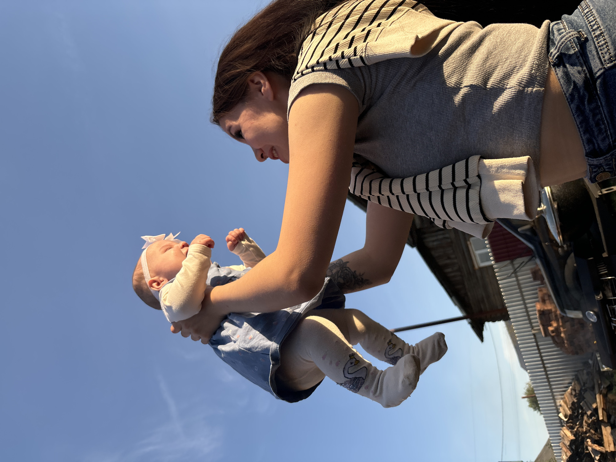 A low-angle shot depicts a woman holding a baby up in the air. The woman has long dark hair and is wearing a gray and white striped shirt, blue jeans, and a striped cardigan. She is looking down at the baby with a gentle expression. The baby is wearing a light blue dress, a white headband with a bow, and white socks with swan designs. The background is a clear blue sky, suggesting it is daytime. A building structure and a vehicle or machinery are visible to the side, indicating an outdoor setting. Given the information provided, it is not possible to confirm that the image location is indeed in Фоки, Russia.